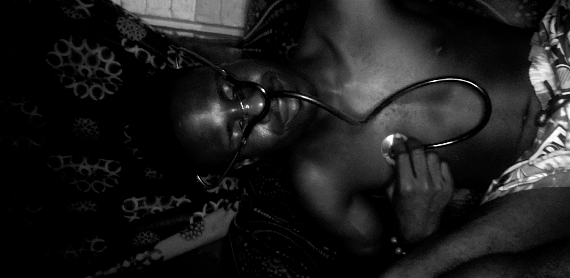 A black and white image shows a person lying on a patterned surface, possibly a bed, using a stethoscope on themselves. The environment is dark, and the location is speculated to be in the city of Narayi, Nigeria. The person's head is angled towards the camera, with the stethoscope earpieces in their ears and the chest piece held to their chest by their hand. The patterns on the surface and the person's clothing add texture to the composition. The overall scene suggests a self-examination or medical check-up.