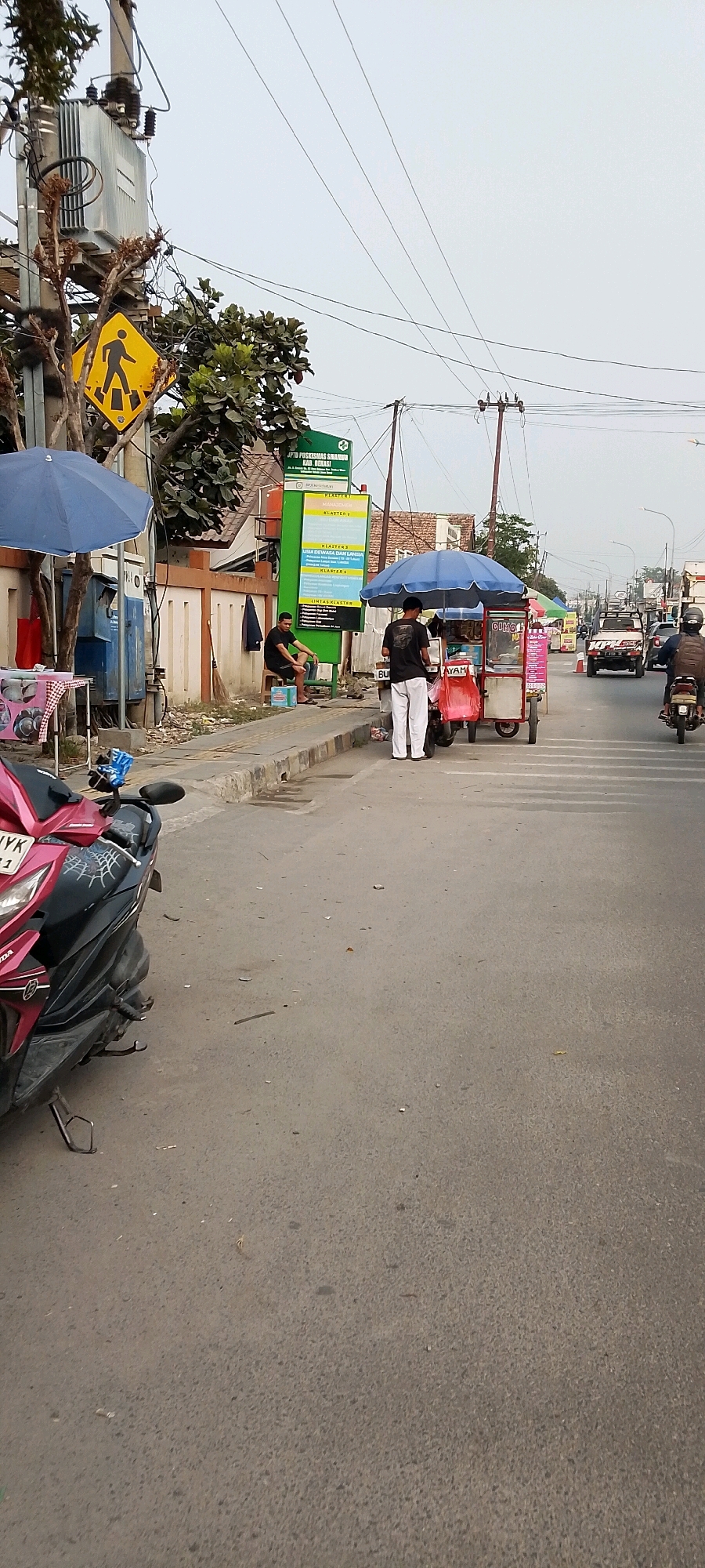 The image depicts a street scene in Sriamur, Indonesia, likely during the daytime with overcast skies. In the foreground, a pink and black motorcycle is parked on the left side of the asphalt road. Further down the street, a food cart with a blue umbrella is set up on the sidewalk, attended by two individuals. One person, wearing white pants and a black shirt, stands behind the cart, while another, wearing a black shirt and pants, sits on a small stool to the left of the cart. A blue umbrella also shades a small table with items on the sidewalk to the left of the food cart.
Behind the food cart, a green sign with white text is mounted on a pole, identifying a local health center, "UPTD PUSKESMAS SRIMUR KAB BEKASI". The sign lists various "CLUSTER" sections, likely related to services or programs offered by the clinic.
The environment includes a sidewalk, a paved road with traffic lanes indicated by painted lines, and electrical wires overhead supported by poles. In the background, buildings and trees are visible, along with other vehicles on the road, including a white pickup truck and motorcycles. A pedestrian crossing sign is prominently displayed on a pole near the transformer on the left side of the image. The overall impression is a busy, functional urban setting.
