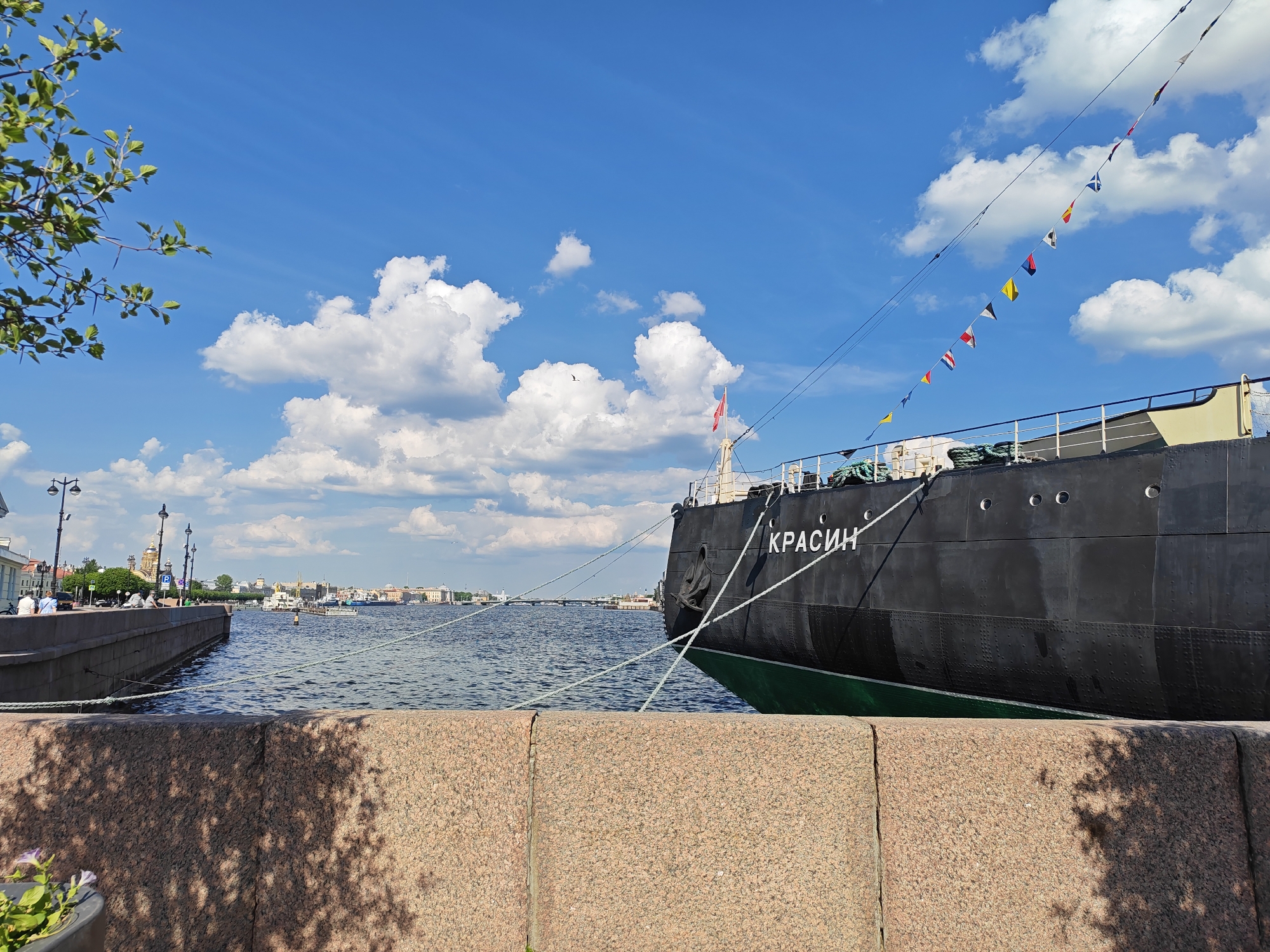This bright daytime image captures a serene scene along a wide waterway in Saint Petersburg, Russia, focusing on the historic icebreaker *Krasin* and the city skyline. In the immediate foreground, a sturdy granite embankment, mottled with dappled shadows from unseen foliage on the left, anchors the composition.
Dominating the right side of the frame is the large, dark grey hull of the ship, identified by the white Cyrillic letters "КРАСИН" (KRASIN) on its bow. A band of green paint is visible along its waterline. The vessel is securely moored to the embankment with several thick white ropes. A decorative string of colorful international maritime signal flags stretches diagonally from the ship's mast upwards to the right, adding a vibrant detail against the sky. A small red flag also flies from the ship near its bow.
The calm, blue water of the waterway occupies the middle ground, reflecting the bright sky and exhibiting gentle ripples. Across the water, the distant cityscape of Saint Petersburg is visible under a clear, bright blue sky adorned with scattered white cumulus clouds. Distinct architectural features, including golden domes and various buildings, line the far bank, alongside streetlights and a blurred "P" parking sign. A bridge spans the waterway further in the distance, and other smaller boats are present on the water. A few indistinct figures of people can be seen on the distant embankment, enjoying the pleasant weather.
The scene suggests a sunny, clear day, likely late morning or early afternoon, with strong natural light creating distinct shadows. The presence of the *Krasin* icebreaker and the architectural style clearly place the setting in Saint Petersburg. The overall mood is peaceful and picturesque.