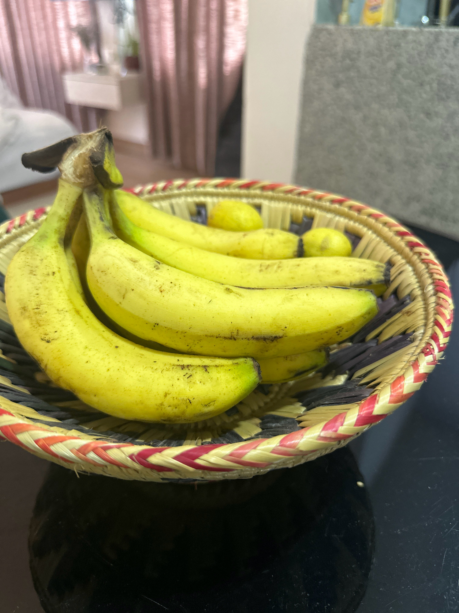 A close-up, eye-level shot shows a woven basket filled with bananas and a few small yellow fruits, possibly lemons. The basket has a red and yellow braided rim. The bananas are mostly yellow with some brown spots, indicating ripeness. The background is blurred, showing hints of a room with pink curtains and furniture. This image was captured in Lalitpur, Nepal.