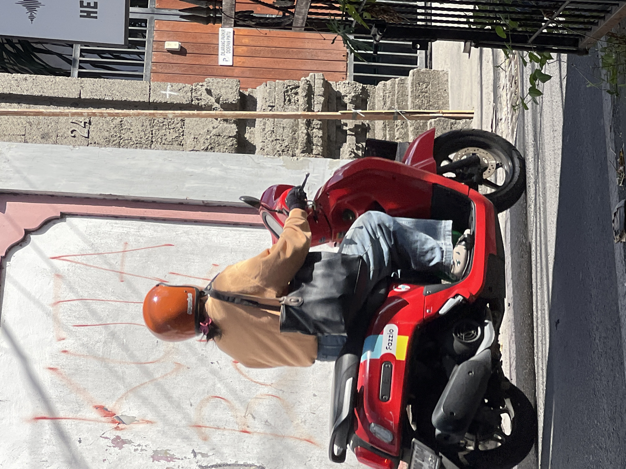 A person wearing an orange helmet and a tan sweater rides a red scooter down a street in Kuta, Indonesia. The scooter has a "Fazzio" logo on its side. The street is paved and lined with buildings. One building has a rough, textured stone facade with the number "24" visible. Another building has a pink and white exterior wall with red graffiti. A black metal gate with green vines growing on it is visible on the right side of the image. A sign on a wooden door reads "DILARANG PARKIR DI DEPAN PINTU," which translates to "NO PARKING IN FRONT OF THE DOOR."