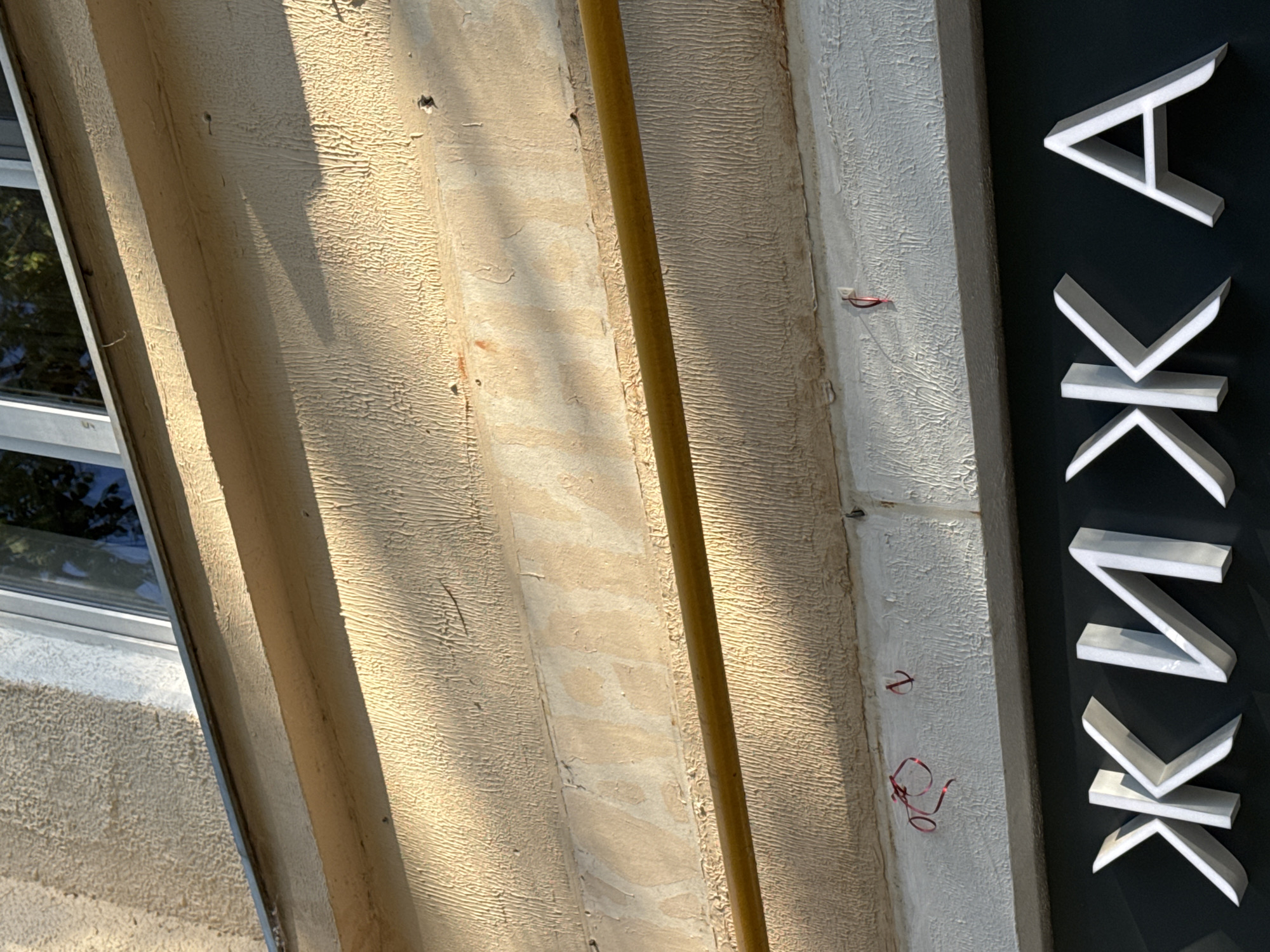 The image depicts a close-up view of a building facade in Moscow, Russia, captured under strong, direct sunlight that creates pronounced shadows and highlights textures.
The upper portion of the frame shows a segment of a window with a light-colored frame, reflecting a dark blue sky or foliage. Below the window, a horizontal band of rough, light-beige stucco features a deeply textured surface, showing signs of wear and age. Further down, another wider band of the same light-beige, heavily textured stucco is visible. Faint, ghosted outlines of removed white Cyrillic letters, possibly spelling "КАФЕ" (CAFE), are discernible within this section, indicating a previous sign. A horizontal yellow gas pipe runs across the mid-section of the beige stucco wall.
Below the gas pipe, the wall transitions to a rough, white-painted stucco. Attached to this white surface are two pieces of thin, tangled red material, possibly string or wire, remnants of a prior fixture. The lower part of the image features a contemporary sign with large, three-dimensional, white Cyrillic letters spelling "ЖИЖА" against a dark, possibly black, background. The letters cast subtle shadows, emphasizing their relief.