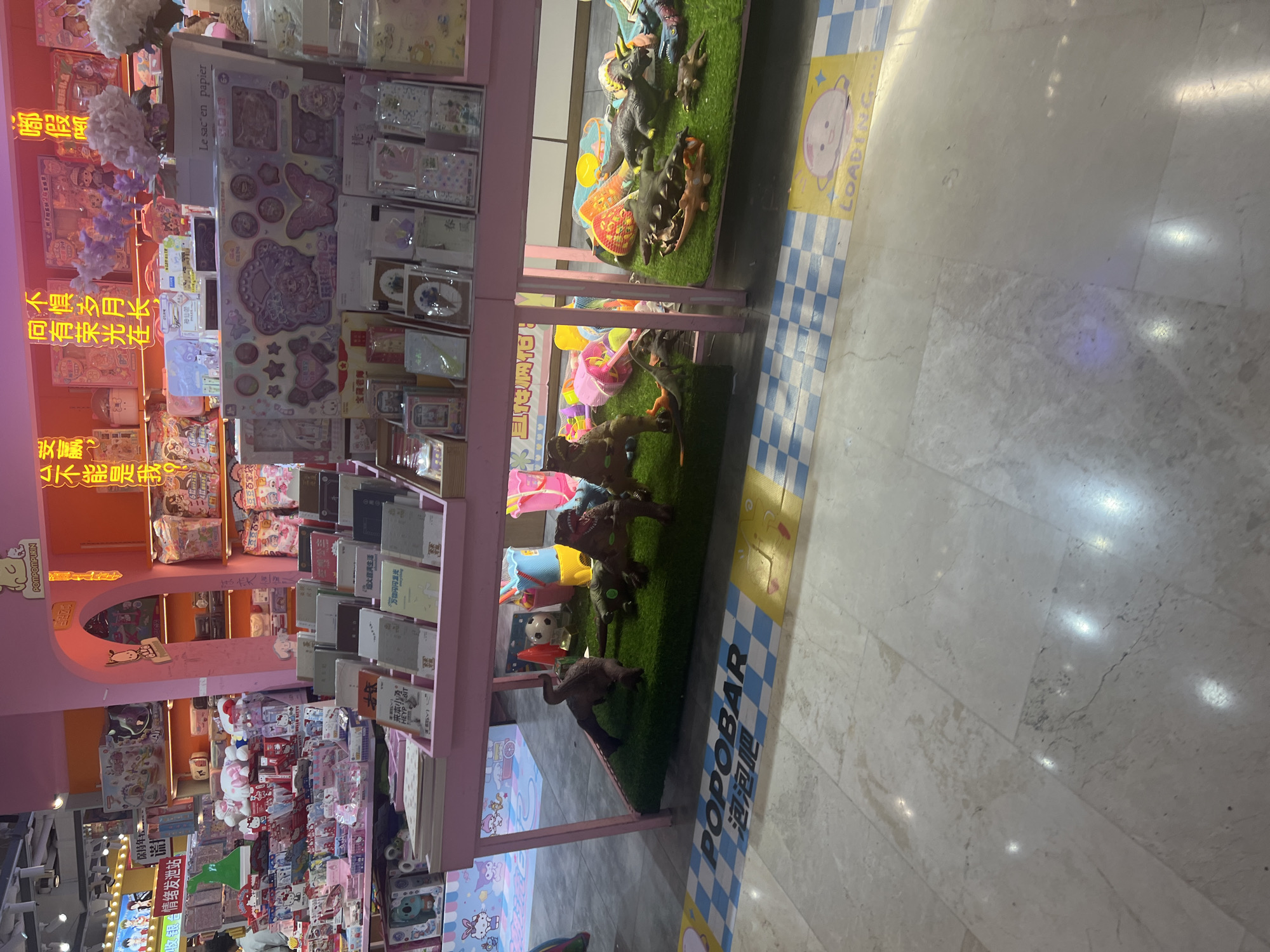 The image shows an indoor retail environment, likely a toy store or a section of a larger department store. The aesthetic is heavily influenced by pastel colors, particularly pink, with bright lighting. In the foreground, a display of dinosaur toys is arranged on artificial grass. These toys are varied in size and species, with several large dinosaur figures and smaller ones scattered among them. Behind the dinosaur display, a pink shelf unit is filled with various items, including books, stationery, and decorative packaging, with some items resembling stickers or craft supplies. The shelves are organized in an appealing manner, suggesting a retail display.
Further back, the store continues with more shelves stocked with a variety of products, including what appear to be plush toys and other children's merchandise. The walls are painted in bright, cheerful colors like pink and orange, with some decorative elements. In the background, there are signs with Chinese text, indicating the location and potentially product information. A prominent sign with the text "POPOBAR" and Chinese characters is visible on the floor in the foreground, suggesting a specific brand or area within the store. The floor is a mix of marble tiles and a checkered pattern. The overall impression is of a vibrant, modern, and child-friendly retail space. While the specific city of Liandu District, China is mentioned as the location, this cannot be confirmed solely from visual cues within the image.