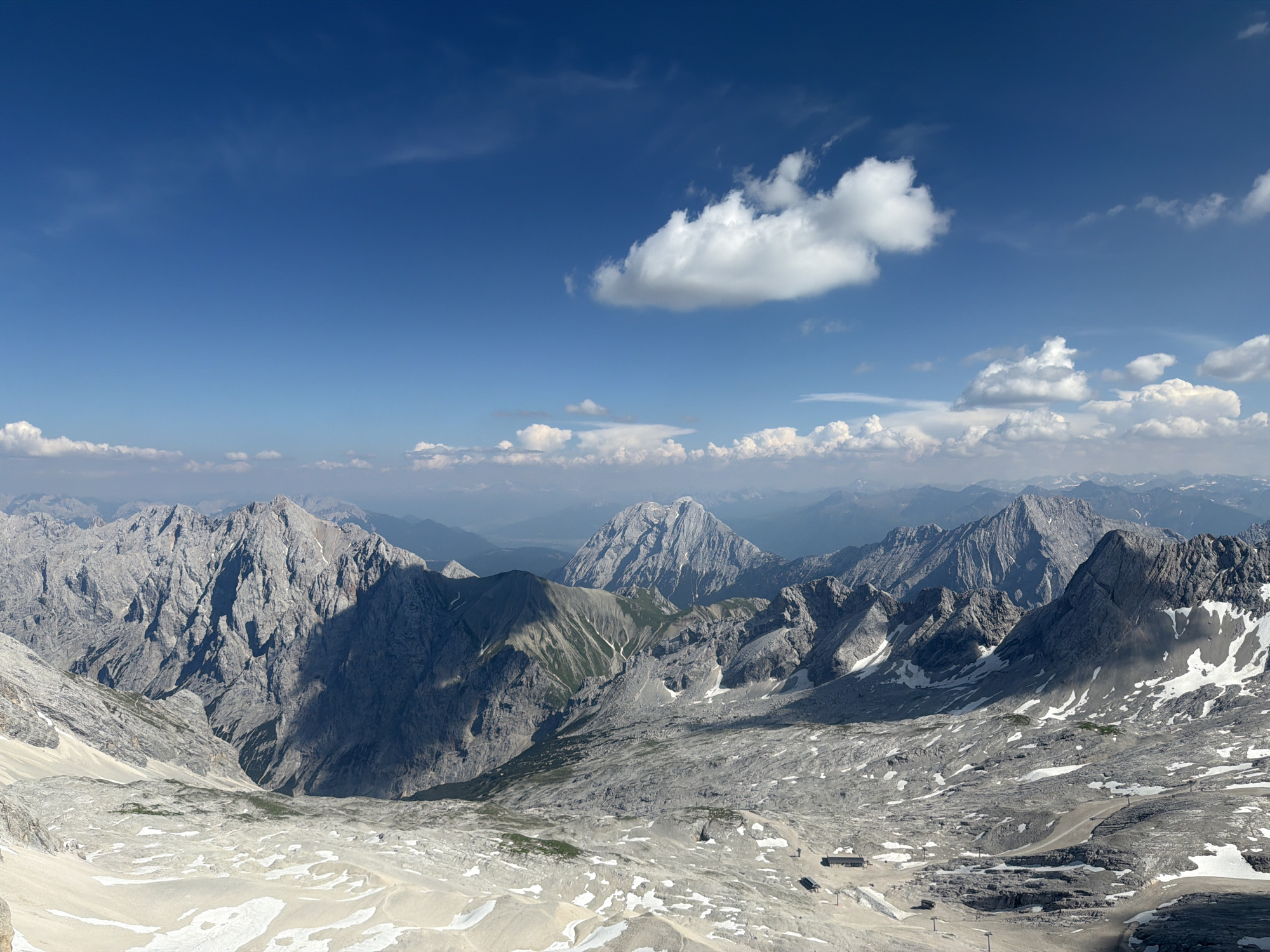 This image captures a breathtaking panoramic view of a rugged mountain landscape under a clear blue sky with scattered white clouds. The foreground features a rocky, snow-dusted terrain, indicative of a high-altitude environment. In the middle ground and background, a series of majestic, grey, rocky mountains stretch as far as the eye can see, their peaks and valleys carved by time and the elements. Some of the mountains have patches of snow on their slopes, suggesting a cool, possibly early summer or late spring season at this elevation.
The scene appears to be a scenic overlook or a vantage point from a high peak, offering a vast vista of the Alps. In the middle distance, a small building complex with a dark roof is visible on the rocky plateau, possibly a mountain hut or a visitor center. Several ski lifts or cable car towers are also subtly present, hinting at the accessibility of this remote location for tourism or recreation. The lighting suggests it is daytime, with the sun casting shadows that accentuate the rugged topography of the mountains. The weather appears to be fair and sunny, with a few fluffy white clouds dotting the deep blue sky, creating a serene and awe-inspiring atmosphere. The overall impression is one of immense natural beauty, solitude, and the grandeur of alpine scenery. While no people are clearly discernible, the presence of infrastructure suggests human activity in the area. The location context of Garmisch-Partenkirchen, Germany, places this scene in the Bavarian Alps.