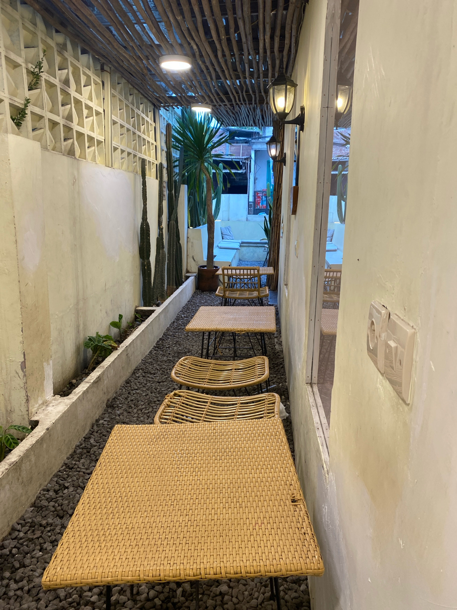 An outdoor seating area is depicted with a narrow walkway lined with gravel and a concrete planter filled with small plants. Tall cactus plants are positioned along the wall on the left. The walkway leads to several small tables and chairs made of woven material. The tables are square or rectangular with woven tops, and the chairs have woven seats and backs. Overhead, a rustic wooden beam ceiling is visible, with two circular lights embedded in it and two traditional wall-mounted lanterns. The area appears to be part of a cafe or restaurant, with glimpses of more seating and structures through a large window or opening on the right. The walls are a light, creamy color. An electrical outlet is visible on the wall to the right.