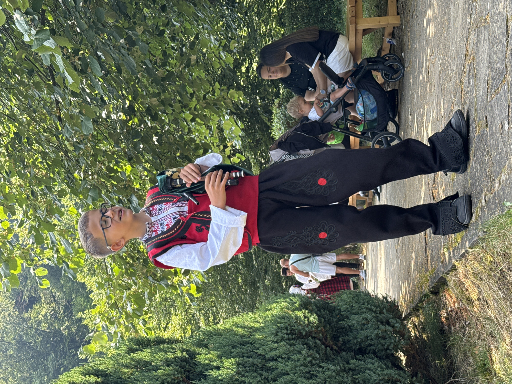 In Ribaritsa, Bulgaria, a young boy in traditional attire, featuring a red vest with intricate embroidery over a white shirt and loose black pants with red floral accents, plays the bagpipes. He stands on a paved path lined with lush green foliage and trees, suggesting an outdoor event or festival. In the background, other individuals are visible; a family sits on a wooden bench, with a woman, a child in a stroller, and another woman present. Further in the background, a few more people are seen walking, some in traditional clothing. The scene is bathed in sunlight, highlighting the vibrant colors of the boy's outfit and the surrounding greenery.