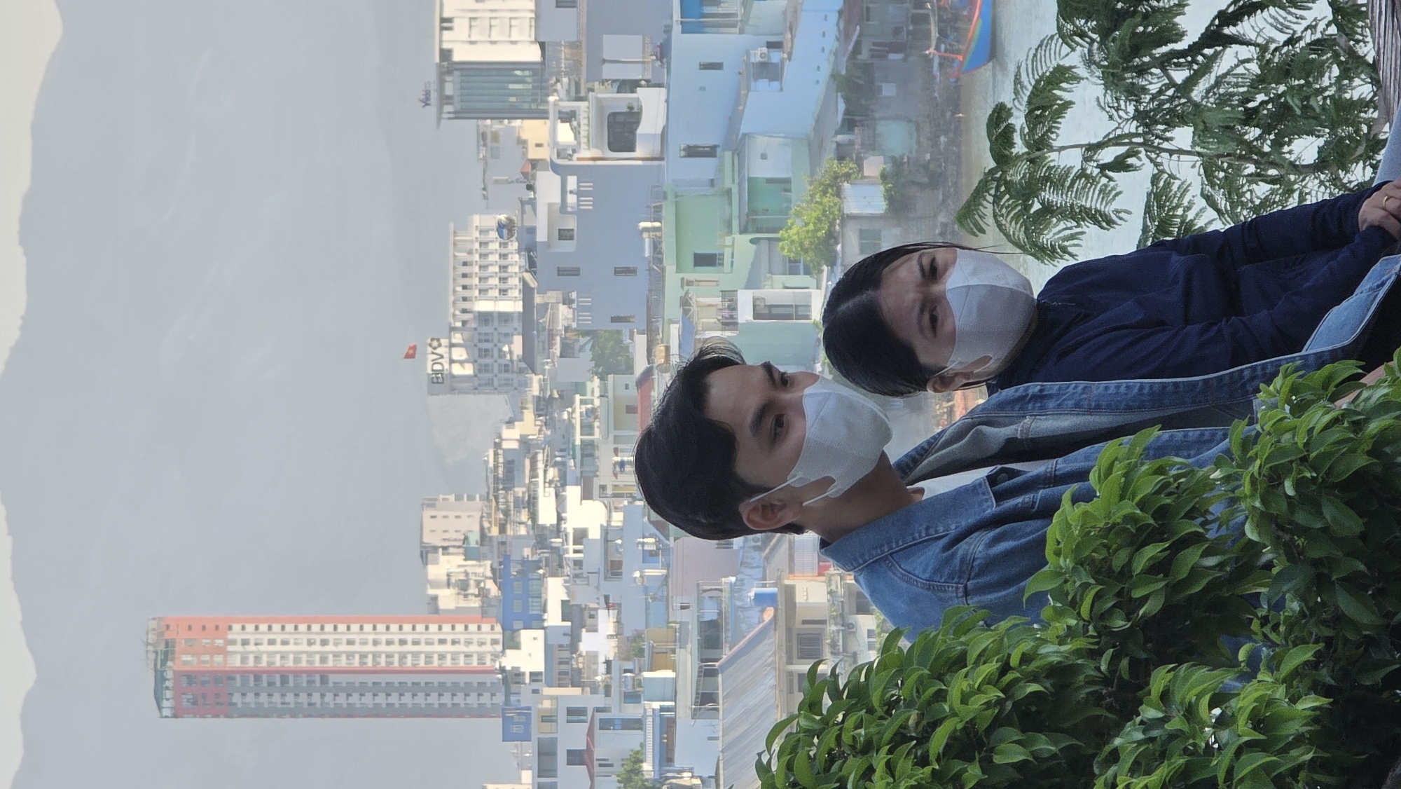 Two individuals wearing face masks are positioned in the foreground, while a dense urban landscape stretches out behind them. The city, identified as Nha Trang, Vietnam, is characterized by its diverse architectural styles and varied building heights. The sea is visible in the upper background, marked by boats and the transition to the horizon. The foreground contains lush greenery, partially obscuring the lower portions of the subjects.