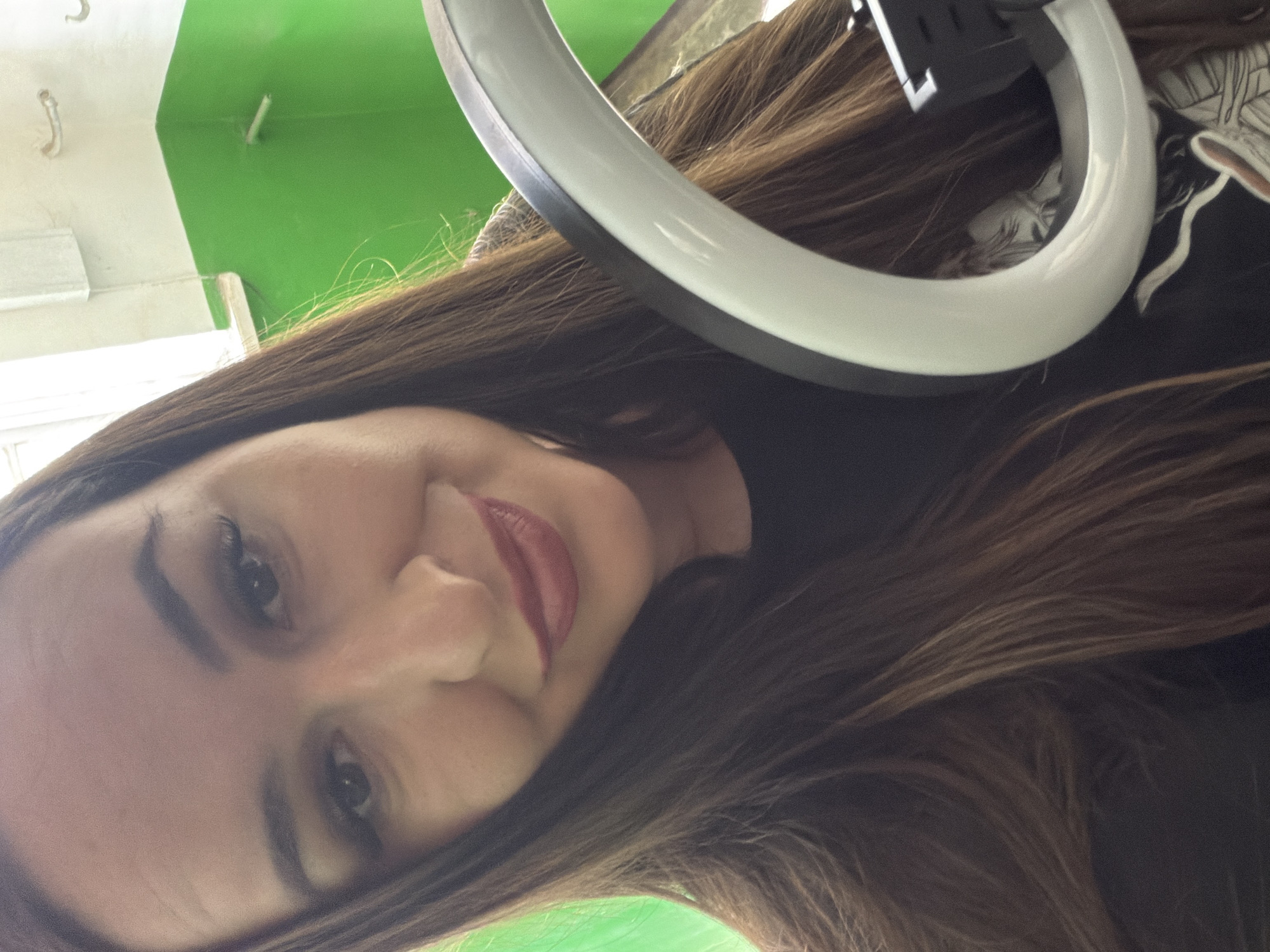 A woman is taking a selfie. She has long, dark brown hair and is wearing makeup. She is holding a ring light up to her face. The background appears to be an indoor space with green and white walls, possibly a hallway or room.