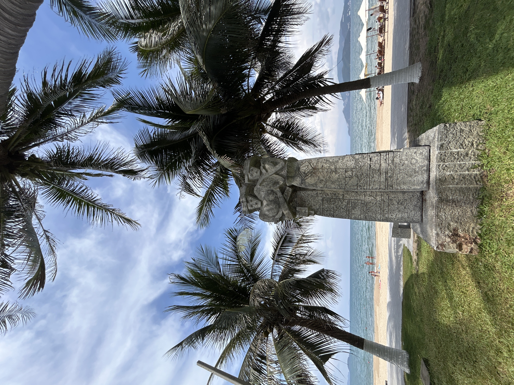 The image depicts a beach scene in Nha Trang, Vietnam. The foreground features a large stone sculpture positioned between palm trees. The sculpture appears to be a monument or decorative element, featuring carved details. Beyond the sculpture is a view of the beach, where people are visible. The beach is lined with palm trees, and further away there are sun loungers and umbrellas, indicating a resort or recreational area. The sky above is blue with scattered clouds. Green grass is also visible on the right of the frame.
