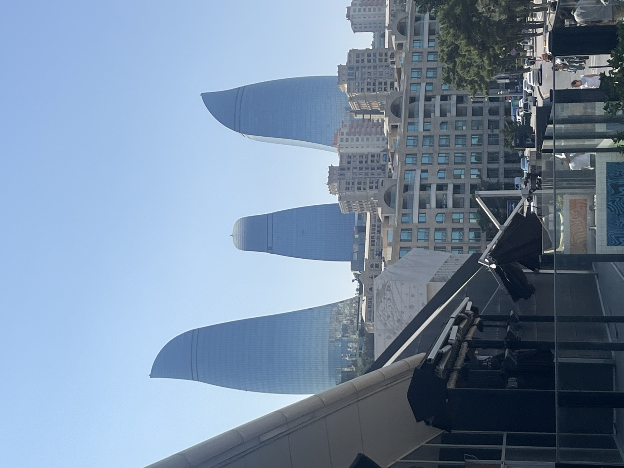 The image captures a daytime, slightly elevated view of the Flame Towers in Baku, Azerbaijan, with a modern building in the foreground. The three Flame Towers, characterized by their flame-like, curved architectural design and glass facades, dominate the background. They are situated in the city and appear to reflect the sky. In front of the towers, a mid-rise building with multiple windows and balconies is visible.
In the immediate foreground, the corner of a contemporary building with a dark facade and a cantilevered section is present. This structure features large windows, a dark roof or overhang, and metal support beams. Below this, a glass barrier encloses an area, beyond which a street scene unfolds. Vehicles, including cars and a truck, are traveling on a road lined with trees. Several individuals are walking on the sidewalk. The sky is clear and blue, suggesting fair weather.