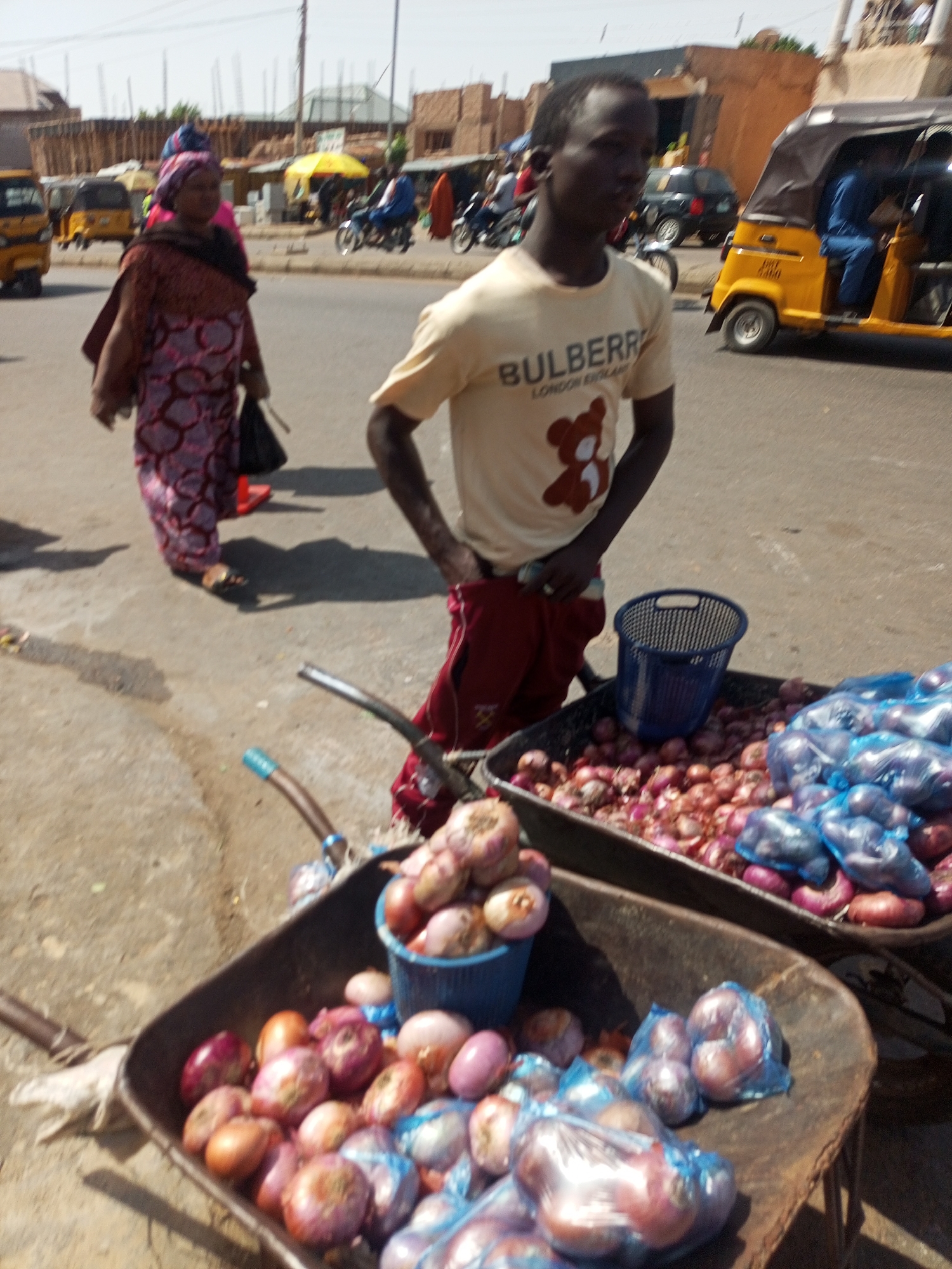A medium shot captures a vibrant street scene, likely a market or commercial area, under bright daylight. In the foreground, a young man, possibly a vendor, stands beside two metal wheelbarrows filled with red and white onions. Some onions are loose, while others are pre-packaged in small blue plastic bags, and a blue plastic basket sits empty in one of the wheelbarrows. The man is wearing a light-colored t-shirt with "BULBERRY LONDON EST. 1824" and a brown bear logo printed on it, paired with red trousers.
In the mid-ground, a woman walks away from the camera, carrying a black bag. She is dressed in a patterned, reddish-brown and pink garment with a head covering. Her feet are visible, wearing sandals. The street surface appears to be asphalt, with some dirt or unpaved areas to the side.
The background reveals a bustling environment with other people, motorcycles, and yellow motor-rickshaws (locally known as "Keke Napep," with the text visible on one vehicle). Buildings constructed of light-colored bricks or concrete are visible, some appearing unfinished. The bright sun casts clear shadows, indicating midday conditions. The overall impression is one of everyday street commerce in a warm climate.