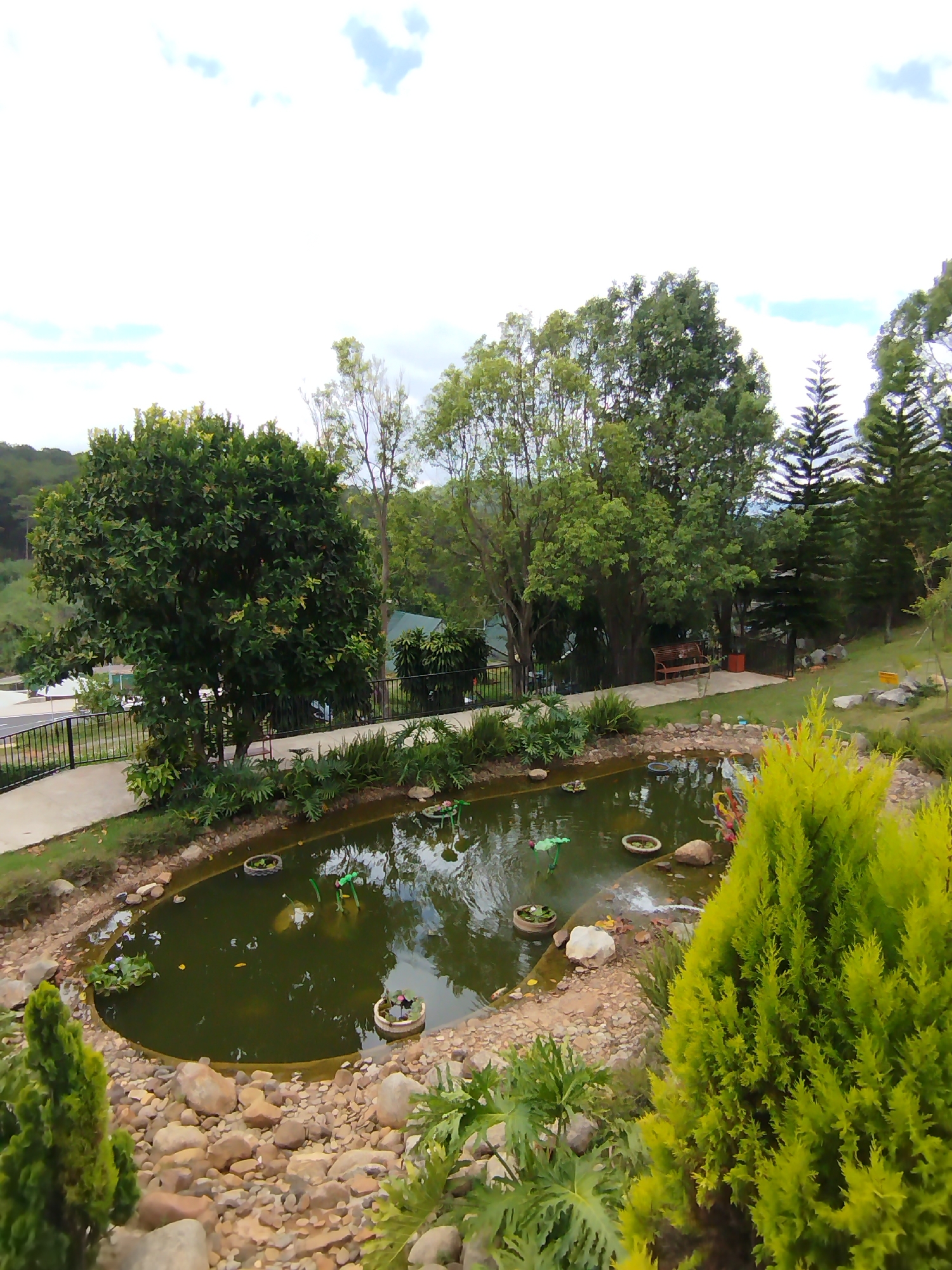 A photograph captures a high-angle view of a small pond in Da Lat, Vietnam, surrounded by greenery. The pond contains floating decorations and possibly small fountains. Various trees and shrubs frame the pond, with a walking path visible along one side. The area around the pond is decorated with rocks. In the background, a park bench can be seen among the trees. The sky is partly cloudy.