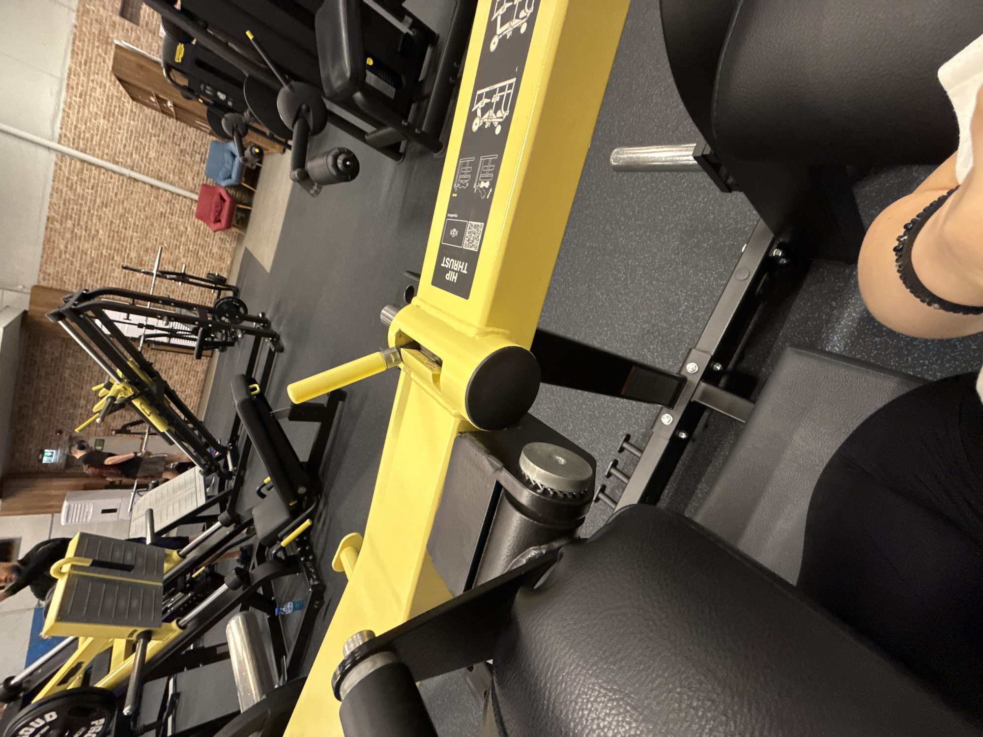 The image captures a slightly tilted view of the interior of a gym in Sofia, Bulgaria. In the immediate foreground, a person is partially visible on the right side of the frame, seated on a black padded bench of a yellow gym machine. Their right arm, adorned with a black elastic bracelet or scrunchie, is visible along with part of their black attire.
The main subject is a prominent yellow hip thrust machine, which dominates the central and right portions of the frame. It features black padded supports and a silver weight pin. On the yellow frame of the machine, clear instructions and branding are visible. The text "HIP THRUST" is prominently displayed vertically, along with illustrative diagrams demonstrating its use and a QR code. Below the diagrams, the brand name "cybex" is faintly discernible.
In the mid-ground and background, various other gym equipment is visible. These include leg press machines, weight racks, and possibly other strength training apparatus, many also featuring yellow and black elements. Several other individuals are present in the background, appearing to be engaged in workouts or moving between machines. The gym floor is a dark gray with speckles, and the walls are a mix of light-colored brick and plain white or light gray. A blue water bottle is visible on the floor near some equipment in the background. The lighting suggests an indoor setting, likely well-illuminated by artificial lights.