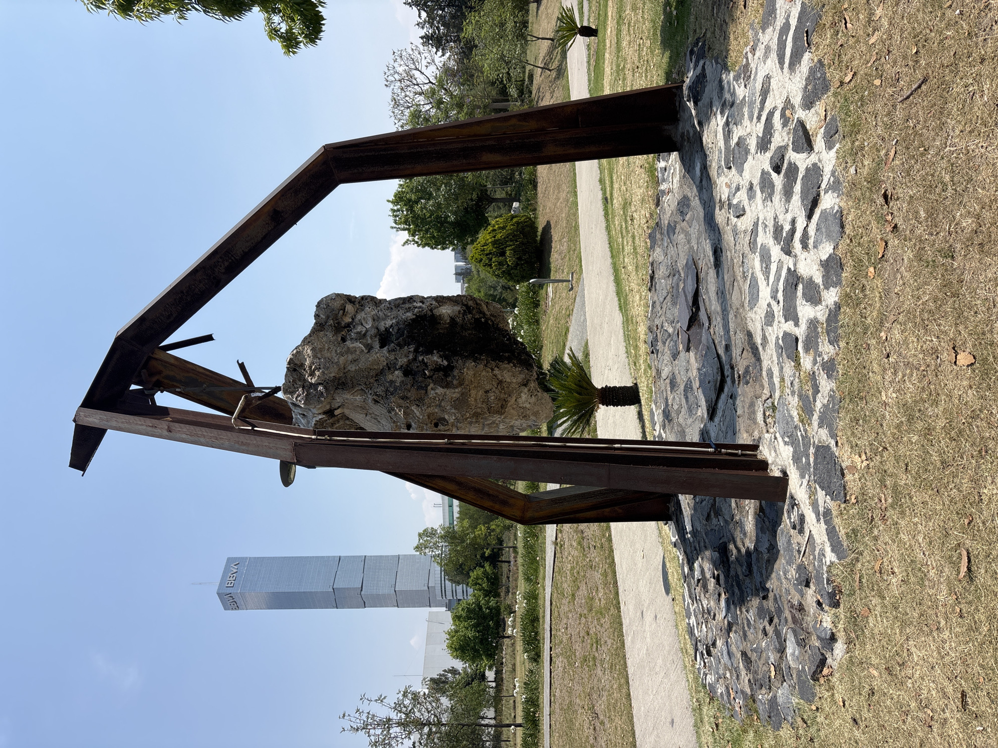 A horizontal shot depicts a sculpture composed of a large boulder suspended within a rusted metal framework, set against the backdrop of a park and the BBVA Tower in San Bernardino Tlaxcalancingo, Mexico. The boulder appears weathered and partially covered with dark patches. The metal framework consists of angular supports and a horizontal beam that suspends the boulder. The sculpture rests on a base of black and white stones, with a paved walkway adjacent to it. The BBVA Tower is a prominent rectangular skyscraper with BBVA branding. The surrounding environment features green trees and a clear, blue sky.