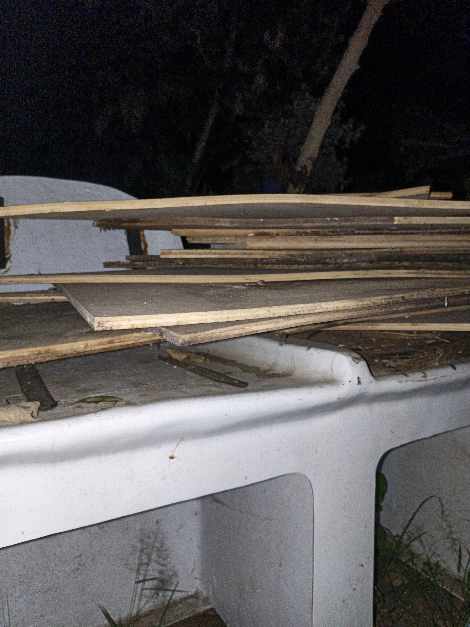 The image displays a pile of wooden planks stacked on a white, curved structure. The planks are of varying sizes and appear weathered, with some showing signs of damage or decay. The white structure looks like it could be part of a boat or a similar vessel, with a hollowed-out section visible. The background is dark, suggesting nighttime, with blurry trees and a trunk of a tree visible. There are no people or animals clearly depicted in the scene. The overall impression is one of disarray or storage of materials in an outdoor setting. The location in Cikal, Indonesia is not discernible from the image itself.