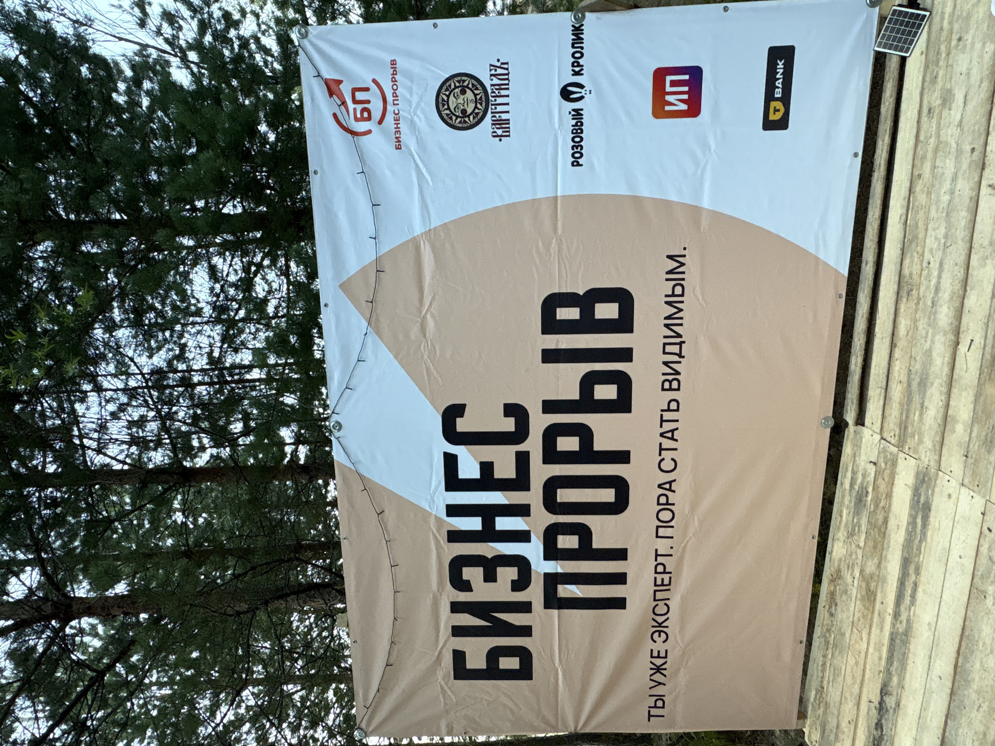 The image shows a large banner displayed outdoors, likely in a natural setting with pine trees visible in the background. The banner is white with a large, light brown diagonal section on the left. Prominently displayed in large, bold black letters on the brown section are the words "БИЗНЕС ПРОРЫВ", which translates to "Business Breakthrough". Below this, in smaller black text, is the phrase "ТЫ УЖЕ ЭКСПЕРТ. ПОРА СТАТЬ ВИДИМЫМ.", meaning "You are already an expert. It's time to become visible."
On the white section of the banner, there are several logos and text elements. In the upper right corner, there is a red circular logo with the letters "БП" inside, accompanied by the text "БИЗНЕС ПРОРЫВ" below it. Further down, there is a circular logo with a sun-like design and some stylized text. Below that, the text "РОЗОВЫЙ КРОЛИК" (Pink Rabbit) is visible with a small rabbit icon. Towards the bottom right, there is a red square with "ИП" (IP, likely an abbreviation for Individual Entrepreneur) inside, and below that, a black rectangular logo with a yellow "T" and the word "BANK".
The banner is supported by wires strung across the top, and it appears to be attached to wooden structures below. The ground is made of wooden planks. There is a small solar panel visible in the lower right corner of the image. The overall scene suggests an outdoor event or promotion. The text elements and logos indicate that this is related to business development and entrepreneurship, possibly a seminar, conference, or marketing campaign. The location is identified as Kokkorevo, Russia.