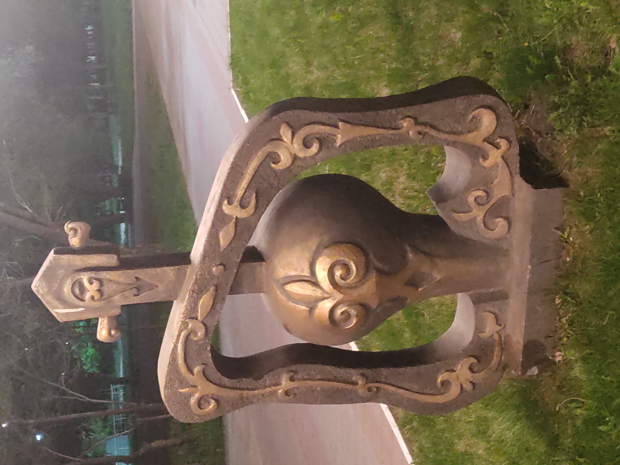 The image depicts a decorative sculpture of a balalaika, a traditional Russian stringed musical instrument, set outdoors. The sculpture is constructed from a grey, stone-like material with gilded embellishments, and its design includes the instrument's distinctive triangular body and neck. It is positioned on a grassy patch alongside a paved pathway. The background shows glimpses of trees and what appears to be a park or public space. There is a lack of specific contextual details to ascertain the exact location within the city of Unknown, Unknown.