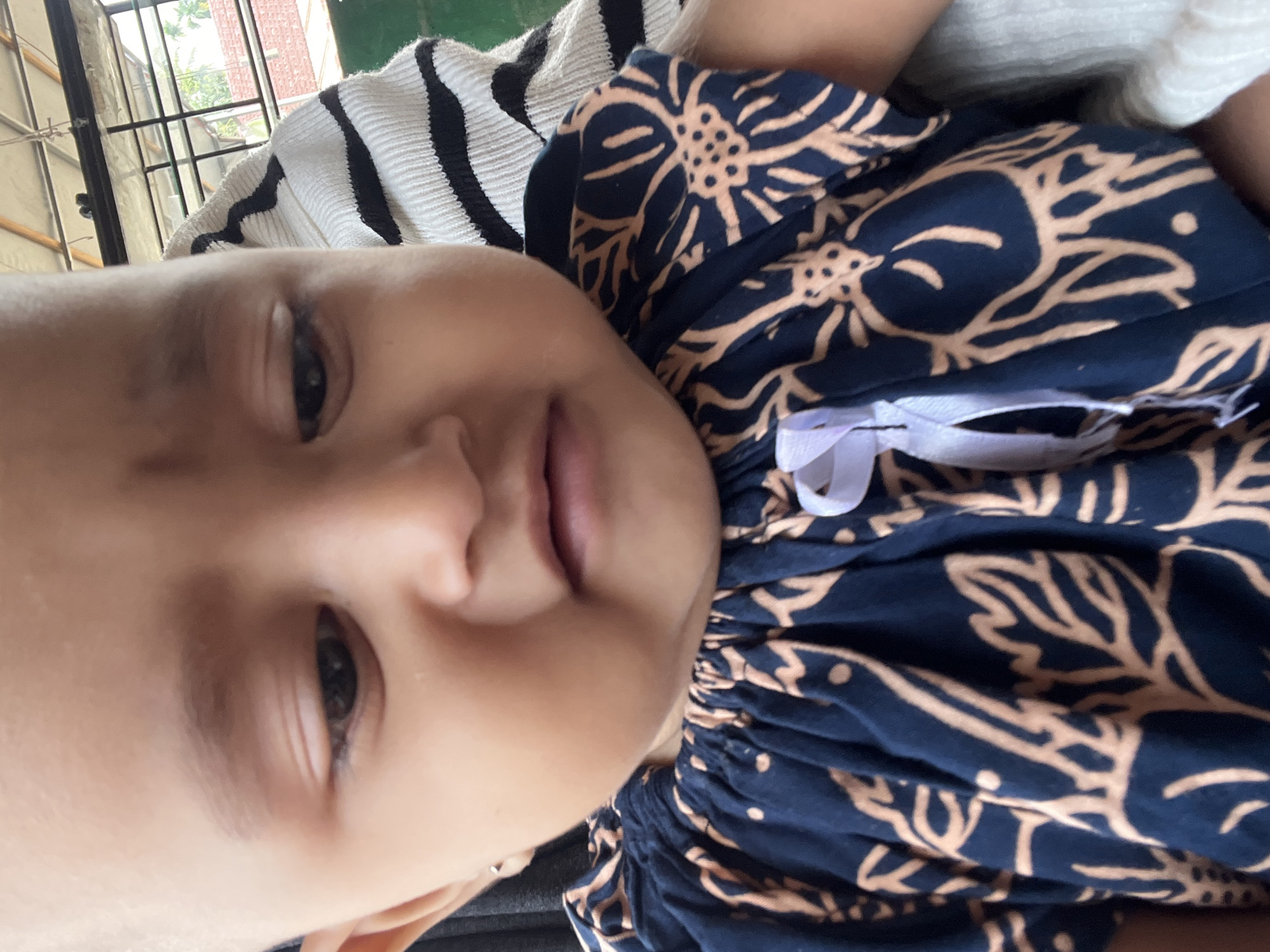 A close-up, downward-angled, outdoor shot captures a baby with a shaved head and fair skin, wearing a dark blue dress with a floral pattern and a white ribbon. The baby's face is partially obscured by the angle, showing one eye, a nose, and a mouth. Behind the baby, a person in a black-and-white striped garment is visible. The background reveals a tiled roof, metal framing, and greenery, suggesting an open-air setting in Seumirah, Indonesia. The overall lighting is natural and appears to be daytime.