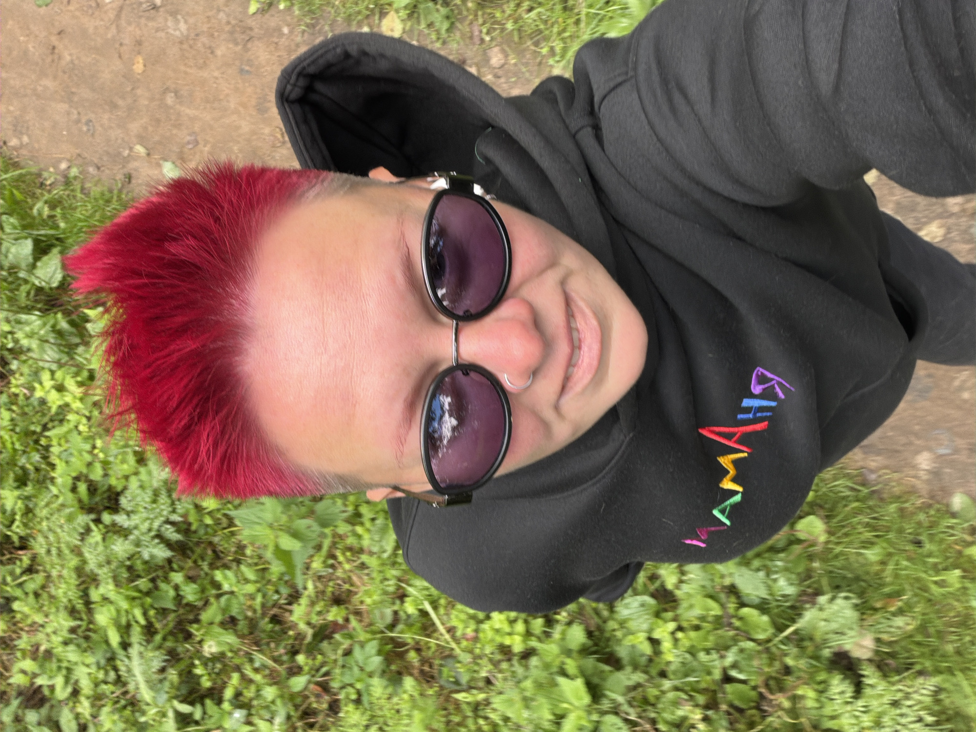 A person with short, spiked, bright red hair is taking a selfie outdoors. They are wearing a black hooded sweatshirt with colorful text on the chest that reads "МАМАЯ". The person is also wearing oval-shaped sunglasses with dark lenses and a silver nose ring. The background consists of green foliage and a dirt path. The image appears to have been taken in the city of Chaikovsky, Russia, as indicated by the text on the sweatshirt.