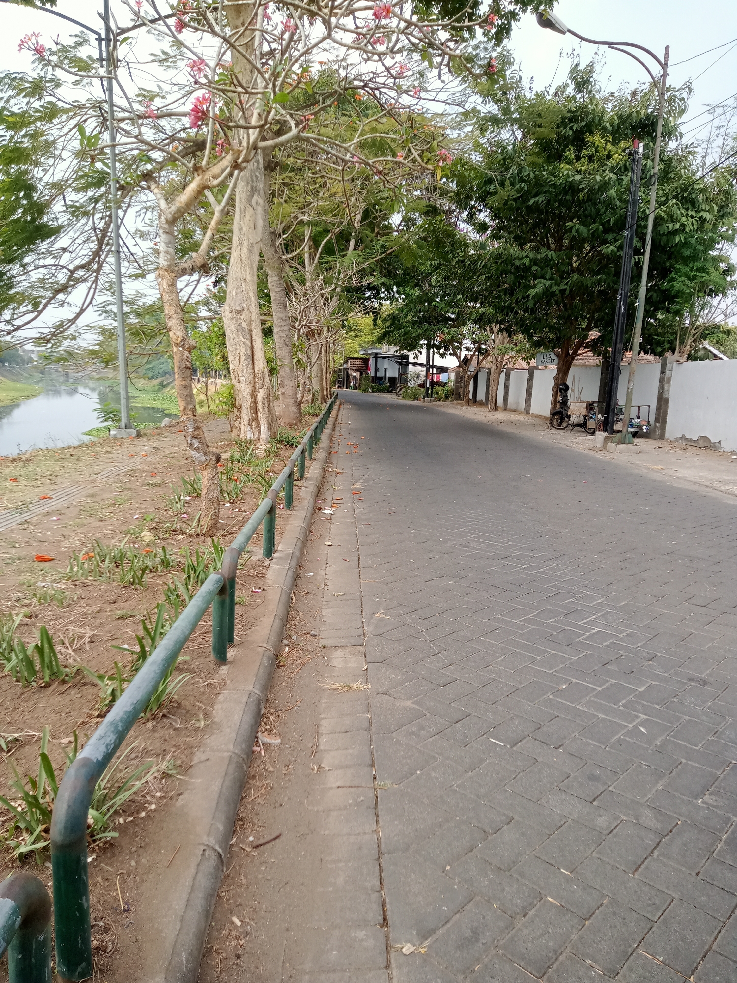The image depicts a street in Tulungagung, Indonesia, bordered by a canal on the left and a wall with buildings on the right. A paved road runs parallel to the canal, with a metal railing separating the walkway from the canal bank. The canal bank is lined with dirt and sparse vegetation, including some green plants and fallen orange flowers. Several trees with slender trunks and light-colored bark stand along the canal bank, some with pink flowers. A few more mature, leafy trees are situated further down the street on the right side, near a white wall. A lamppost with a curved arm is visible behind these trees. In the distance, on the right side of the street, two motorcycles and a cart are parked next to the wall, and a sign that reads "LAS KARBIT" is attached to a pole. The sky is overcast, suggesting a possibly cloudy day. There are no people clearly visible in the image.