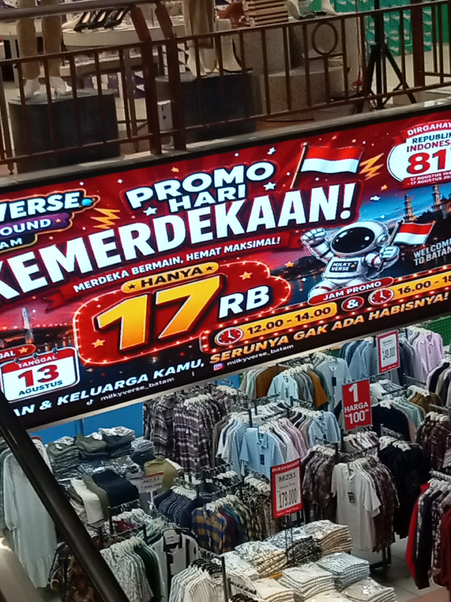 A wide banner advertising a "Promo Hari Kemerdekaan" (Independence Day Promo) is displayed across the upper portion of the image, superimposed over a retail store's clothing racks. The banner features the Indonesian flag, a cartoon astronaut holding the flag, and prominent text promoting deals. Below the banner, rows of folded and hanging clothing are visible, including t-shirts, shirts, and possibly pants. Several price tags are attached to the clothing items. The scene appears to be an indoor shopping environment, likely a clothing store in Batam, Indonesia, given the banner's theme and language. The lighting suggests an indoor setting.