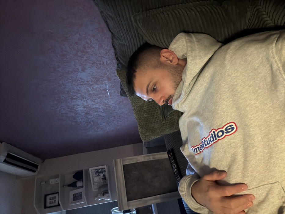 The image displays an indoor residential setting in Sofia, Bulgaria. A male individual, appearing to be in his 20s or 30s, with short dark hair and a trimmed beard, is seen reclining on a dark green, ribbed sofa. He wears a light grey hooded sweatshirt with a red and blue "inetstudios" logo vertically printed on the chest. His head is turned to the right, and his eyes are directed off-camera. His left hand rests on his midsection.
The background features a wall on the right with a distinctive purplish-red textured finish. On the left, a lighter-colored wall holds a white corner shelving unit displaying various decorative items, including framed pictures, two bottles, and a white statuette. Above the shelving unit, a white wall-mounted air conditioning unit is installed. Below the shelves, a framed object, possibly an artwork or mirror with a light-colored frame and dark interior, is visible. A dark-colored remote control rests on the sofa to the subject's left. The room is dimly lit.