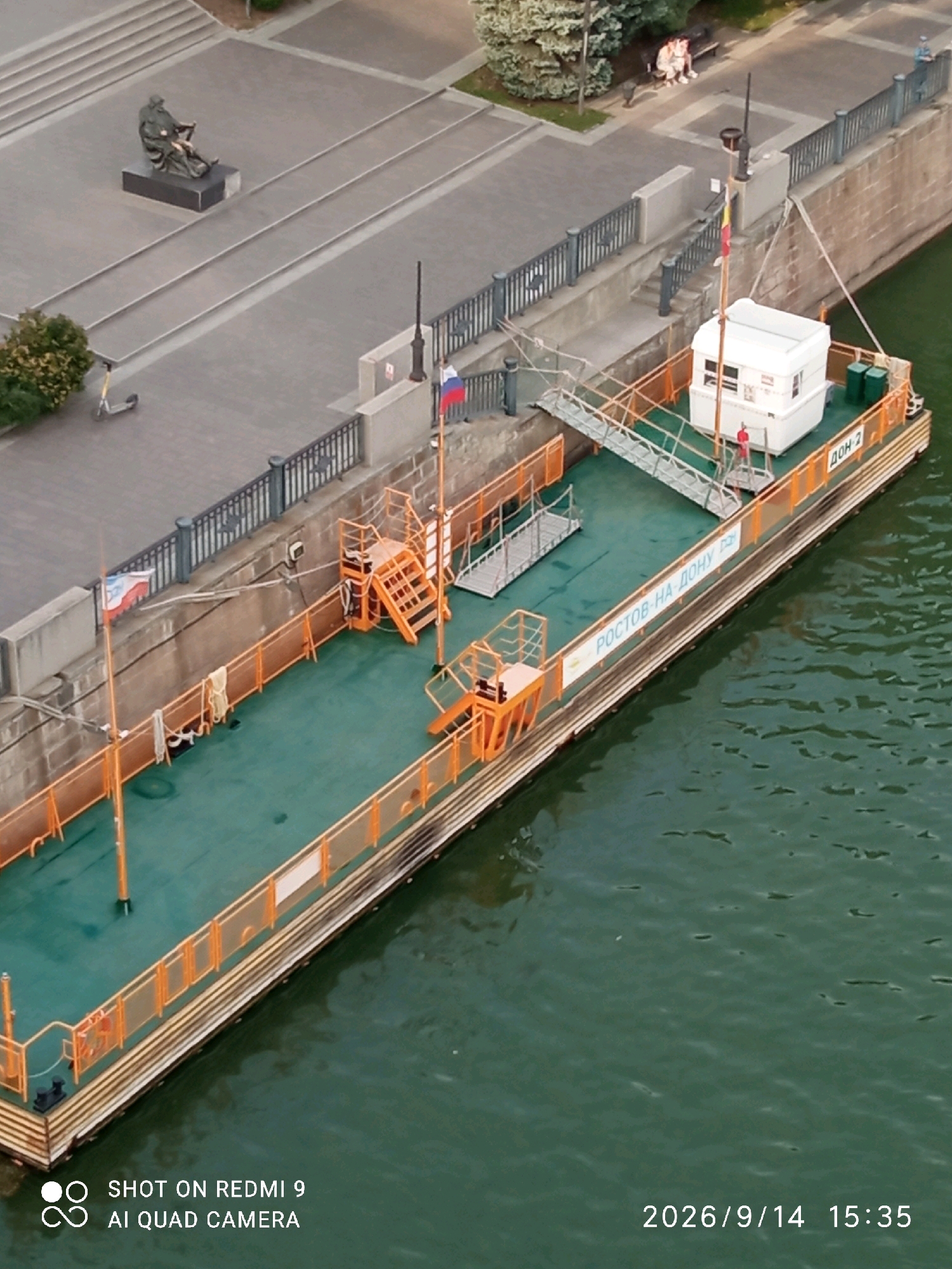 This aerial photograph captures a scene along the Don River embankment in Rostov-on-Don, Russia. The focal point is a long, narrow barge with an orange railing and a green deck, docked alongside a stone embankment. The barge appears to be a passenger ferry or a pleasure craft, indicated by the presence of a small white cabin-like structure on its deck, a ramp providing access, and orange safety equipment. Text on the side of the barge reads "РОСТОВ-НА-ДОНУ" (Rostov-on-Don) and "ДОН-2" (Don-2), further confirming the location and identifying the vessel. The embankment itself features a walkway with a metal railing, a statue of a seated figure, and some sparse vegetation. The water of the Don River is a murky green, with visible ripples on its surface. The image also includes a timestamp in the lower right corner: "2026/9/14 15:35". No people are actively engaged in any specific actions, though two figures are visible on a bench in the background near some trees, and a single figure can be seen near the cabin on the barge.