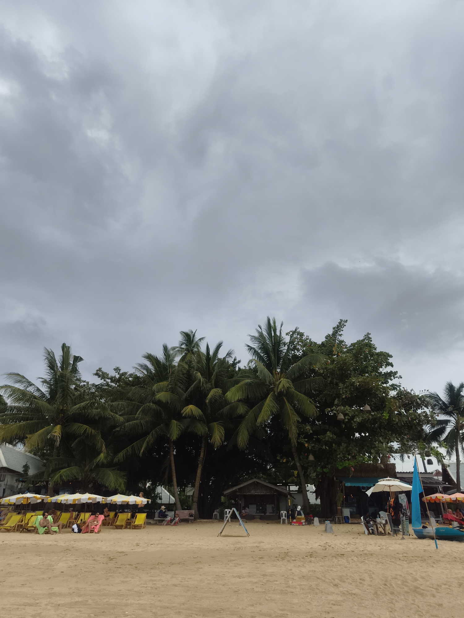 The image shows a beach scene in Baan Mae Nam, Thailand, under an overcast sky. A sandy beach stretches into the foreground. In the middle ground, there are several clusters of beach chairs shaded by striped umbrellas, some of which are occupied by people. Behind the chairs, a line of trees, including palm trees, obscures structures such as beachside businesses. A small building with a thatched roof is visible amongst the trees, and there is a boat near the beach's edge.