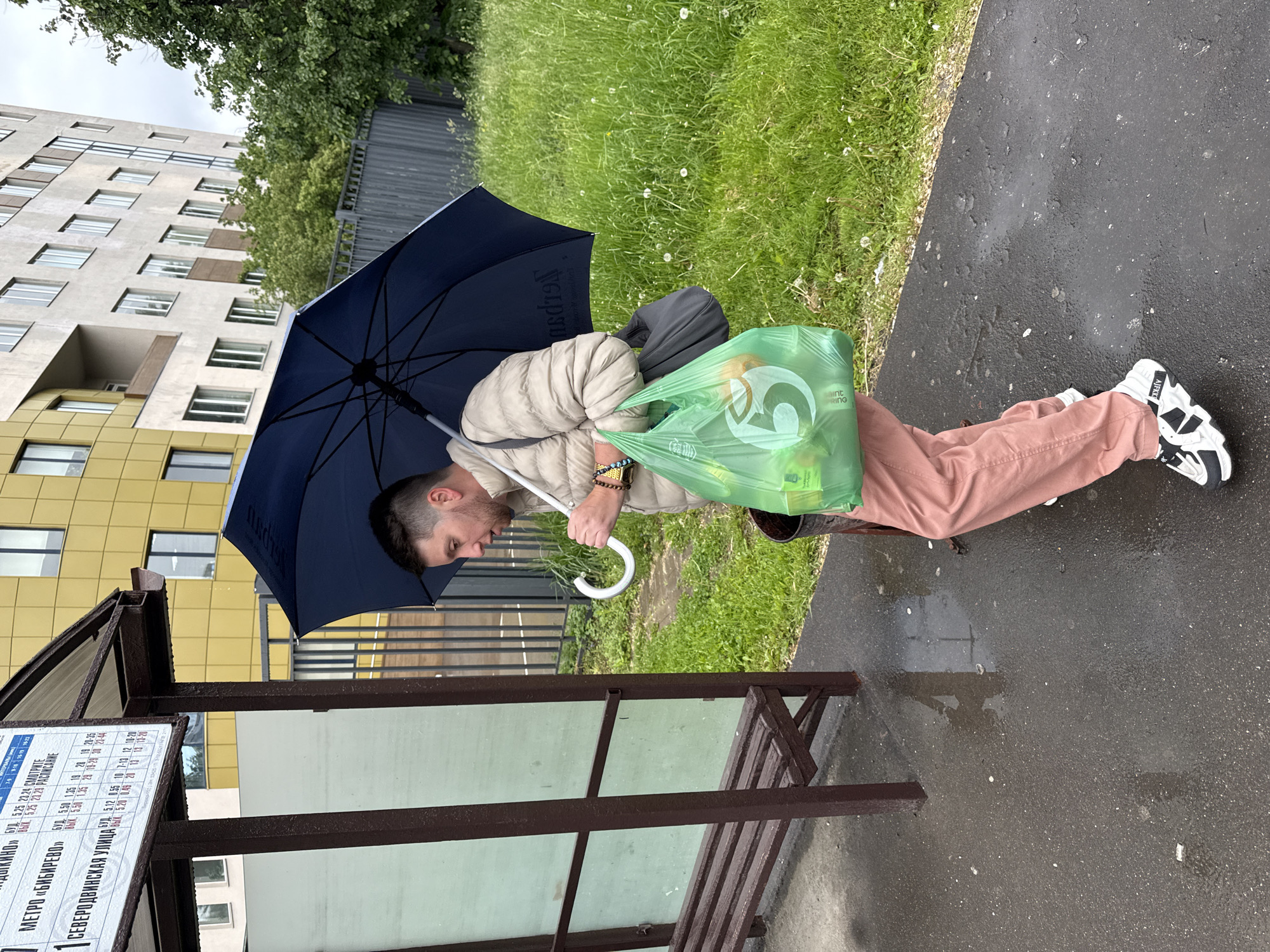 A man stands at a bus stop in Moscow, Russia on a rainy day. He holds a dark blue umbrella over his head and a green shopping bag in his arm. He is wearing a light-colored jacket, pink pants, and white and black sneakers. The bus stop has a timetable with Cyrillic writing including information about bus routes to "Metro Bibirevo" and "Severodvinskaya Street". There is a building visible in the background. The ground is wet from the rain.
