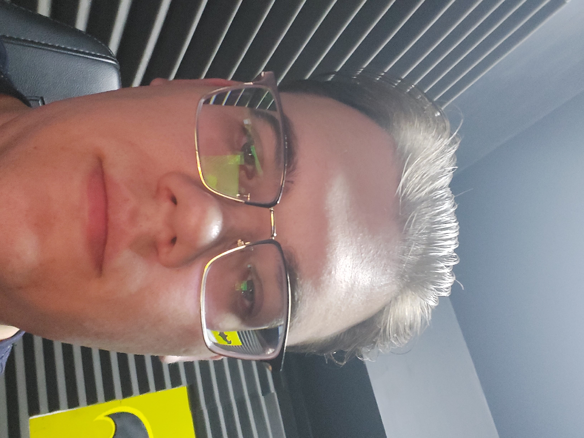 A close-up, slightly angled selfie of a fair-skinned person wearing eyeglasses. The background consists of parallel, dark-colored slats, possibly part of a ceiling or wall structure, and a brighter area suggesting a light source. A yellow object with a black design is visible in the lower left. The person's hair appears light-colored and somewhat disheveled. The reflection in the eyeglasses suggests an interior environment with various colors and shapes. No information regarding Tashkent, Uzbekistan can be obtained from the image.