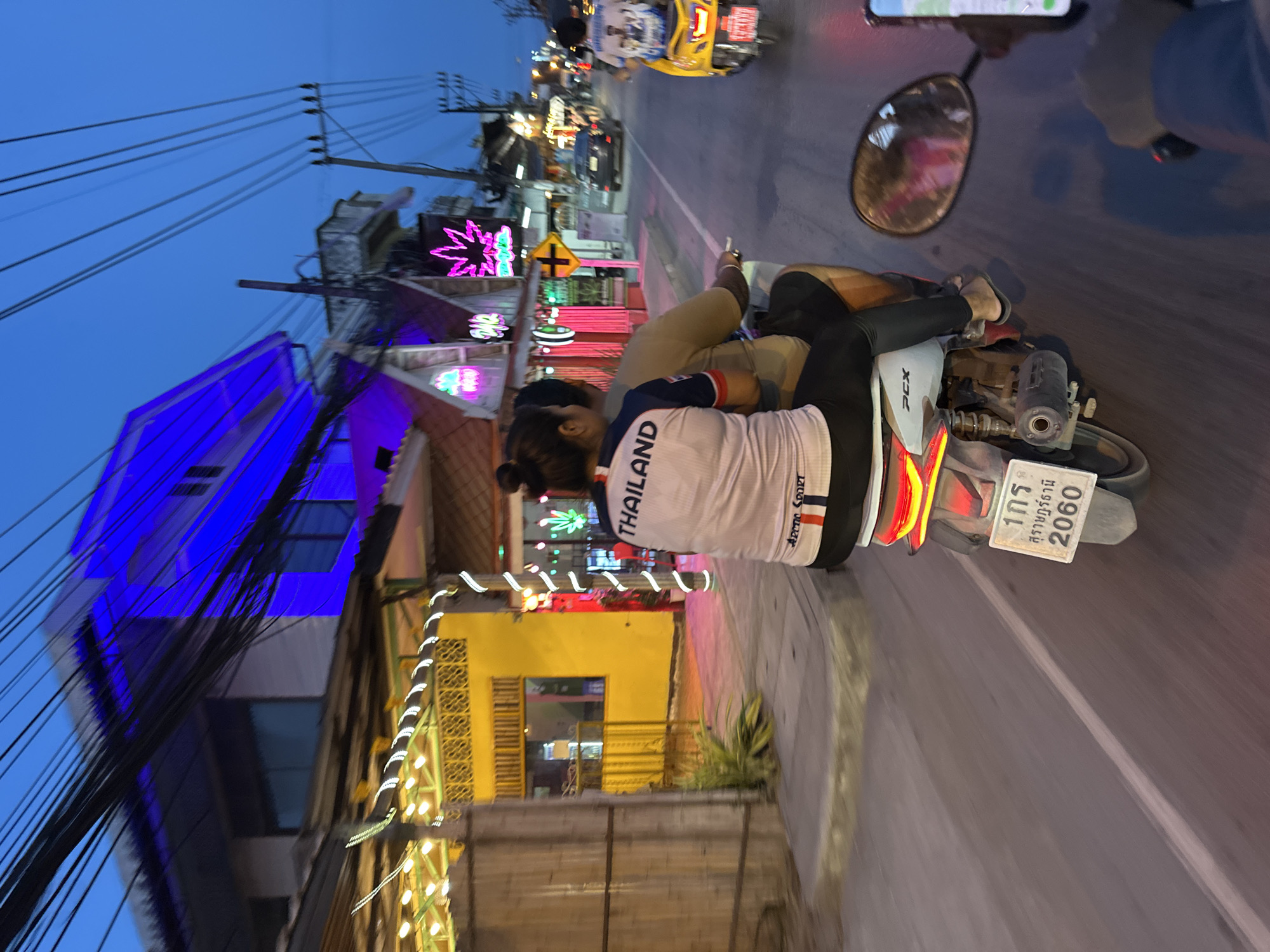 This image depicts a vibrant street scene in Baan Mae Nam, Thailand, at dusk or early night. The sky is a deep blue, and artificial lights illuminate the surroundings. In the foreground, a person is seated as a passenger on a white PCX scooter, viewed from behind. They are wearing a white long-sleeved shirt with "THAILAND" and "ARCTIC SPORT" printed on the back. Another person's leg and foot, wearing a flip-flop, are partially visible, suggesting they are driving or a front passenger.
The scooter's rear license plate displays Thai script, including "1กร" and "สุราษฎร์ธานี" (Surat Thani), along with the numbers "2060". The street is lined with buildings featuring various colorful neon signs, including bright blue, pink starbursts, and a green stylized cannabis leaf, indicative of a commercial or entertainment area. Numerous black electrical wires crisscross the upper left portion of the image, suspended from utility poles.
The scooter is on an asphalt road with a visible white lane marking. Other vehicles, including a yellow vehicle possibly a tuk-tuk, are blurred in the background, suggesting movement or a bustling environment. A rearview mirror and a phone displaying a map or navigation app are visible on the right, attached to the scooter's handlebars. The overall impression is that of an active evening street scene in a Thai town.