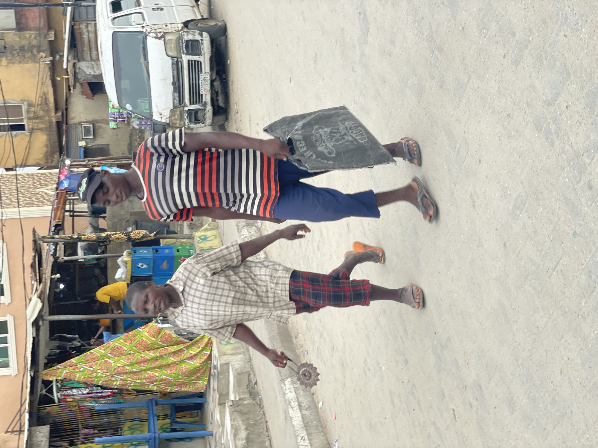 The image depicts a vibrant street scene, likely in an urban or semi-urban area of Ado, Nigeria, during daylight hours. Two men are walking on a paved road, with buildings and a parked vehicle forming the backdrop.
The man on the left wears a black baseball cap, a vertical striped t-shirt (red, black, and white), dark blue shorts, and flip-flops. He carries a large, dark grey sack over his shoulder, which has the word "BAGCO" and other text partially visible on it.
The man on the right wears a light-colored plaid shirt, red and black plaid shorts, and mismatched flip-flops (one orange, one greyish). He holds a metal tool with a serrated, gear-like wheel in his right hand, possibly a sharpener or a hand-grinder. Both men are looking towards the viewer with neutral to slightly smiling expressions.
In the background, a white utility van or mini-bus is parked. Its license plate on the front bumper clearly shows "LAGOS STATE" and "GGU 987 BV." Behind the vehicle and the men, various buildings, some with windows and corrugated metal roofs, line the street. Several blue crates, likely for drinks, and colorful patterned textiles are visible, suggesting nearby shops or market stalls. The bright, even lighting indicates a clear, sunny day. The scene captures daily life and movement on a local street.