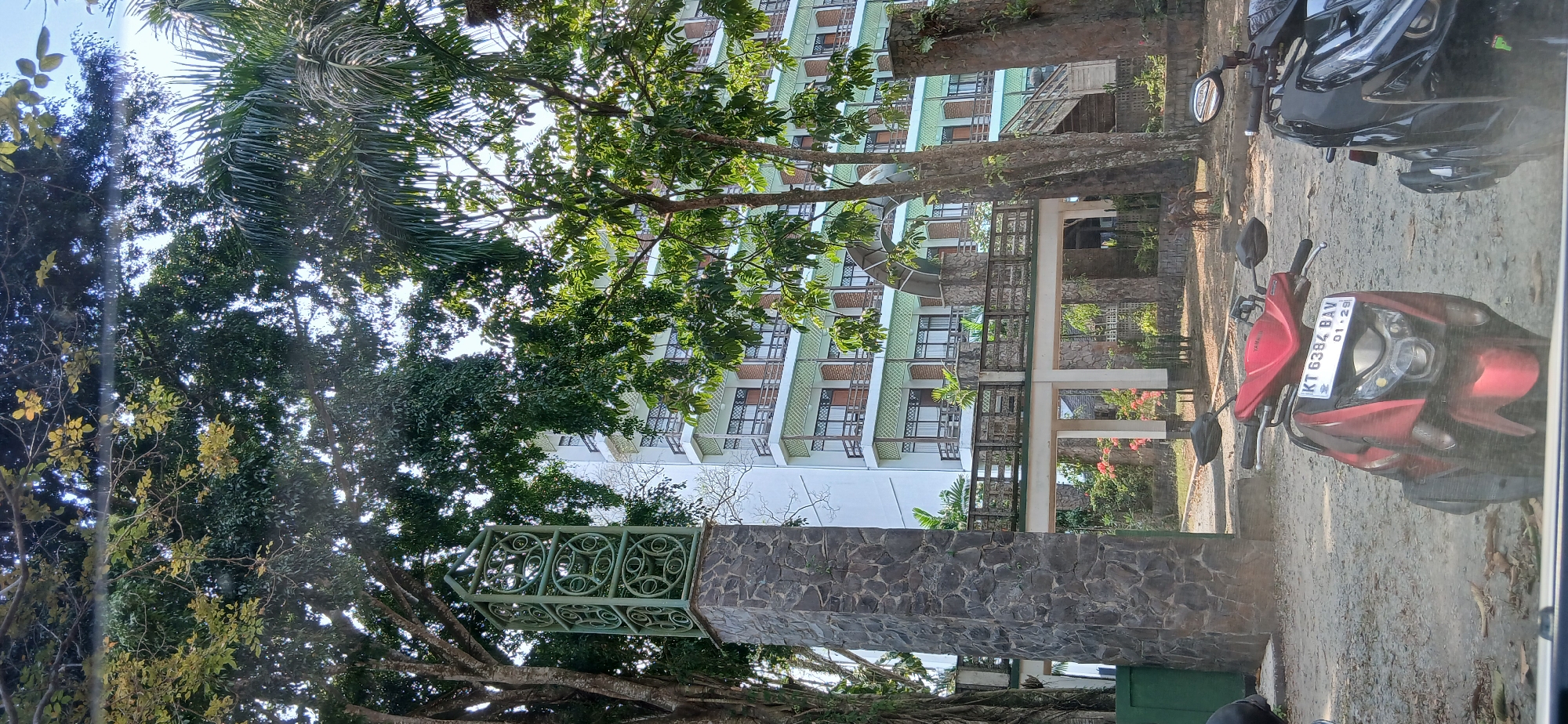 The image depicts a building with multiple stories, possibly a hotel or apartment complex, with balconies and windows visible. In front of the building, there's a decorative stone pillar with a green metal structure on top. Trees and foliage surround the area, with lush greenery and some palm trees. In the foreground, two motorcycles are parked on a dirt and gravel surface. The red motorcycle in front has a license plate that reads "KT 6384 BAV 01-29". The scene appears to be outdoors, under daylight. Based on the license plate format, specifically the "KT" prefix which is common for East Kalimantan, and the overall environment suggesting a tropical climate, this location is likely in the city of Samarinda, Indonesia. There are no people or discernible actions taking place in the image.