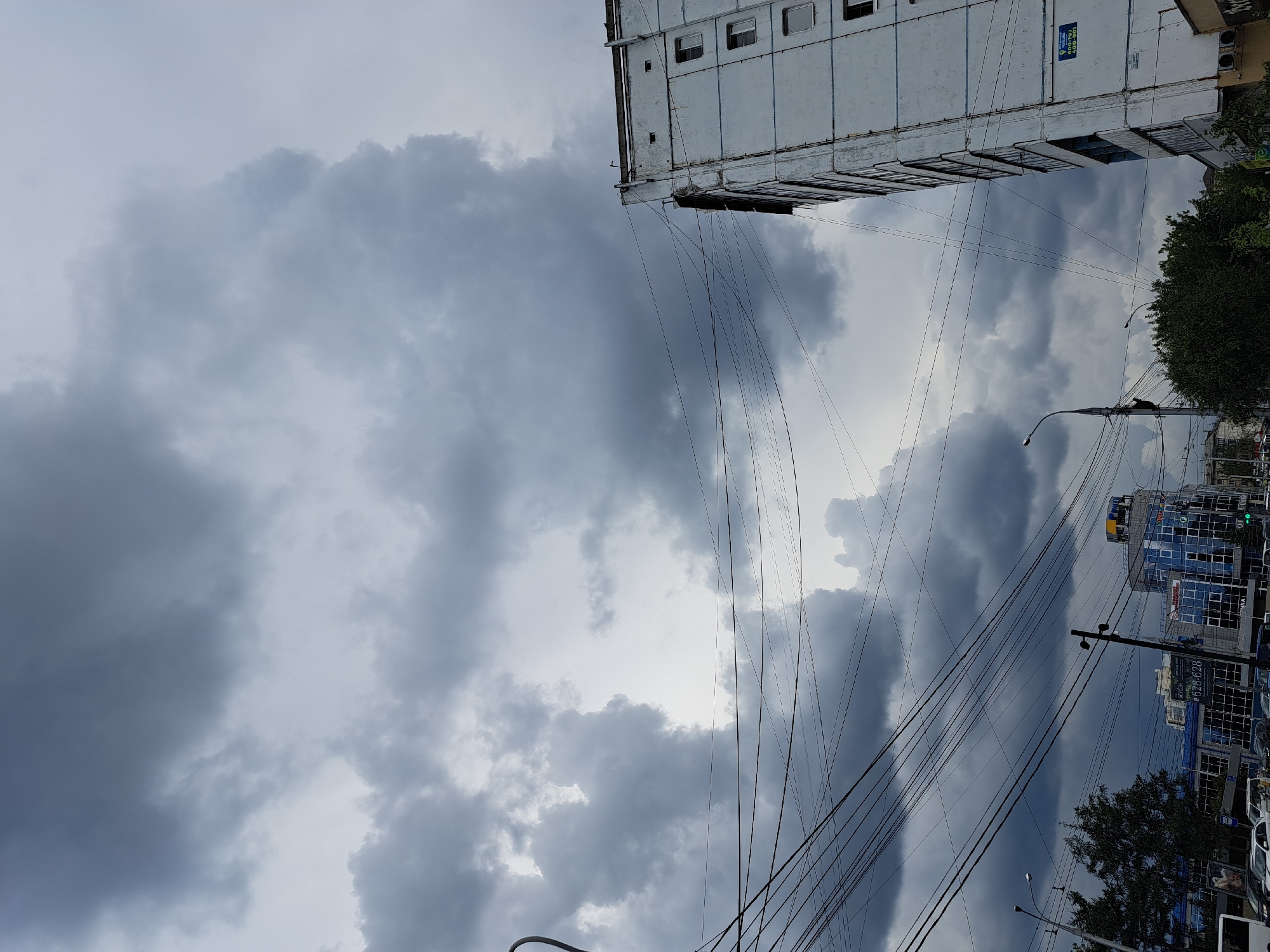 The image shows a cloudy sky above a city street in Irkutsk, Russia. On the right is a tall, white apartment building with multiple windows. In the foreground, a dense network of electrical wires crisscrosses the frame. To the left, trees and other buildings, including a modern glass-fronted structure with signs, are visible. The overall impression is of an overcast day in an urban setting.