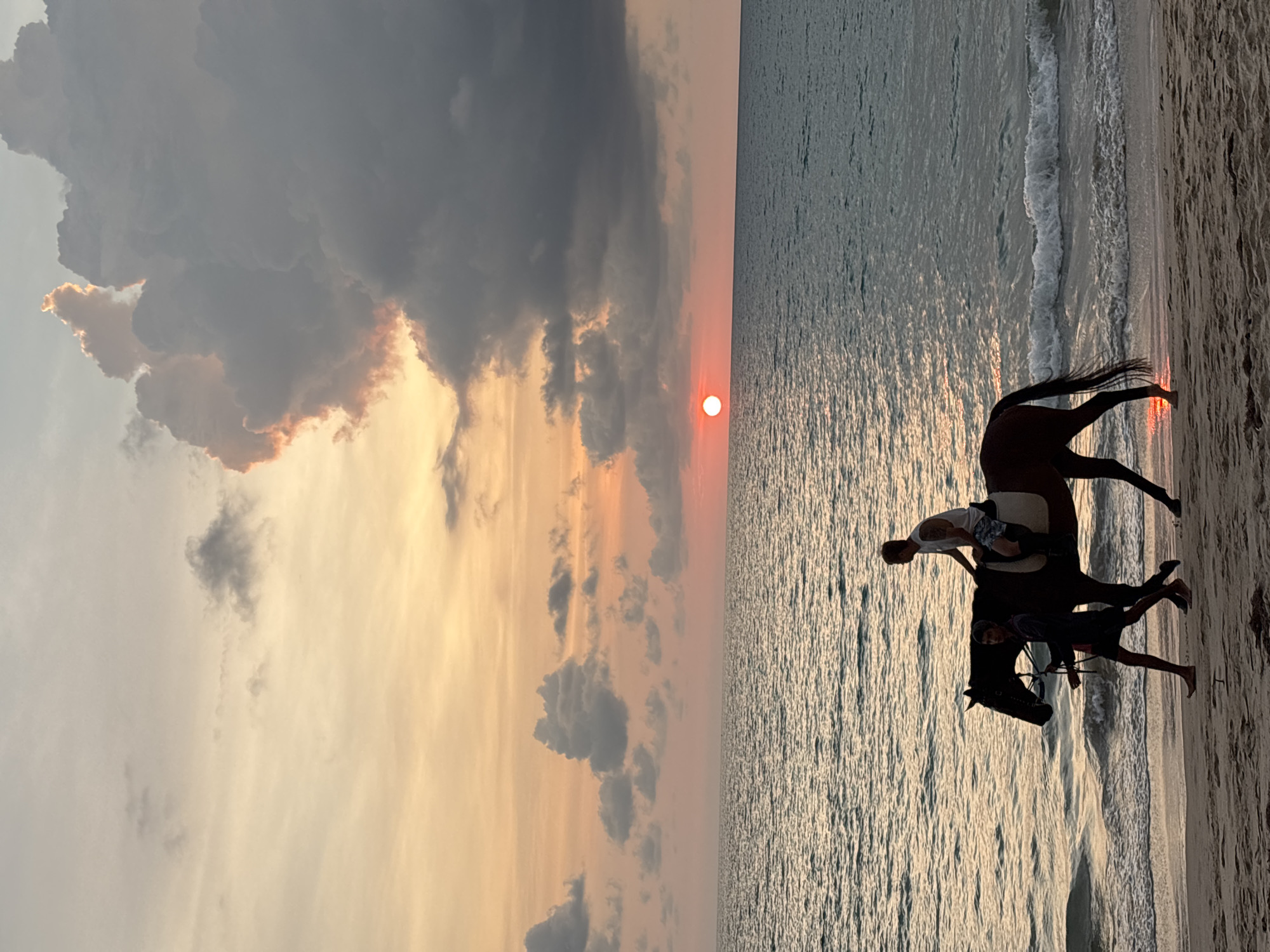 An image captures a serene sunset scene on a beach in Choeng Thale, Thailand. In the foreground, a dark horse is being ridden by one person, while another person walks alongside, appearing to lead the horse near the water's edge. The person riding is wearing a light-colored top, and the person leading is wearing darker attire.
The setting is a picturesque beach with the calm ocean to the right, showing gentle waves breaking on the sandy shore. The water's surface reflects the warm, golden light of the setting sun, creating a shimmering effect.
The most striking feature is the dramatic sky. The sun, a vivid orange-red circle, is positioned low on the horizon where the sky meets the ocean. Above, the sky is filled with a mix of clouds; some are dark and heavy, while others are illuminated from beneath and around the edges by the golden and orange glow of the sunset, casting warm hues across the scene. The overall impression is one of tranquility and natural beauty at dusk. There is no visible text in the image.