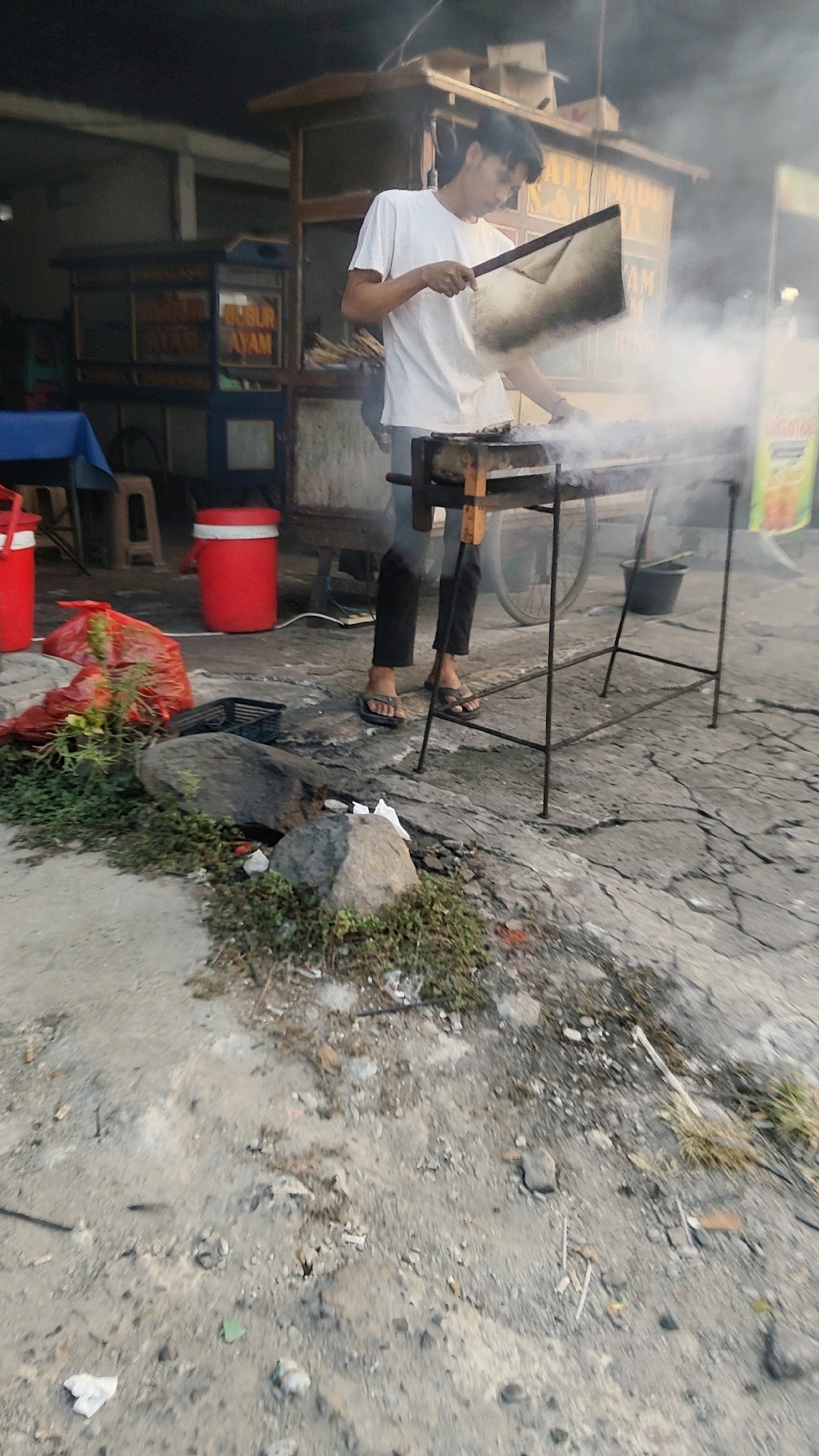 A young man in a white t-shirt and dark pants is grilling outdoors. He is holding a metal fan, seemingly fanning the coals. A grill sits in front of him, with meat cooking on it and smoke rising. In the background, there is a food stall with signage that partially reads "GUR AYAM". Red plastic containers and a red plastic bag are visible on the ground. The ground appears to be a cracked concrete surface with some debris and sparse vegetation. The scene suggests a street food vendor operation in Pasirsari, Indonesia.