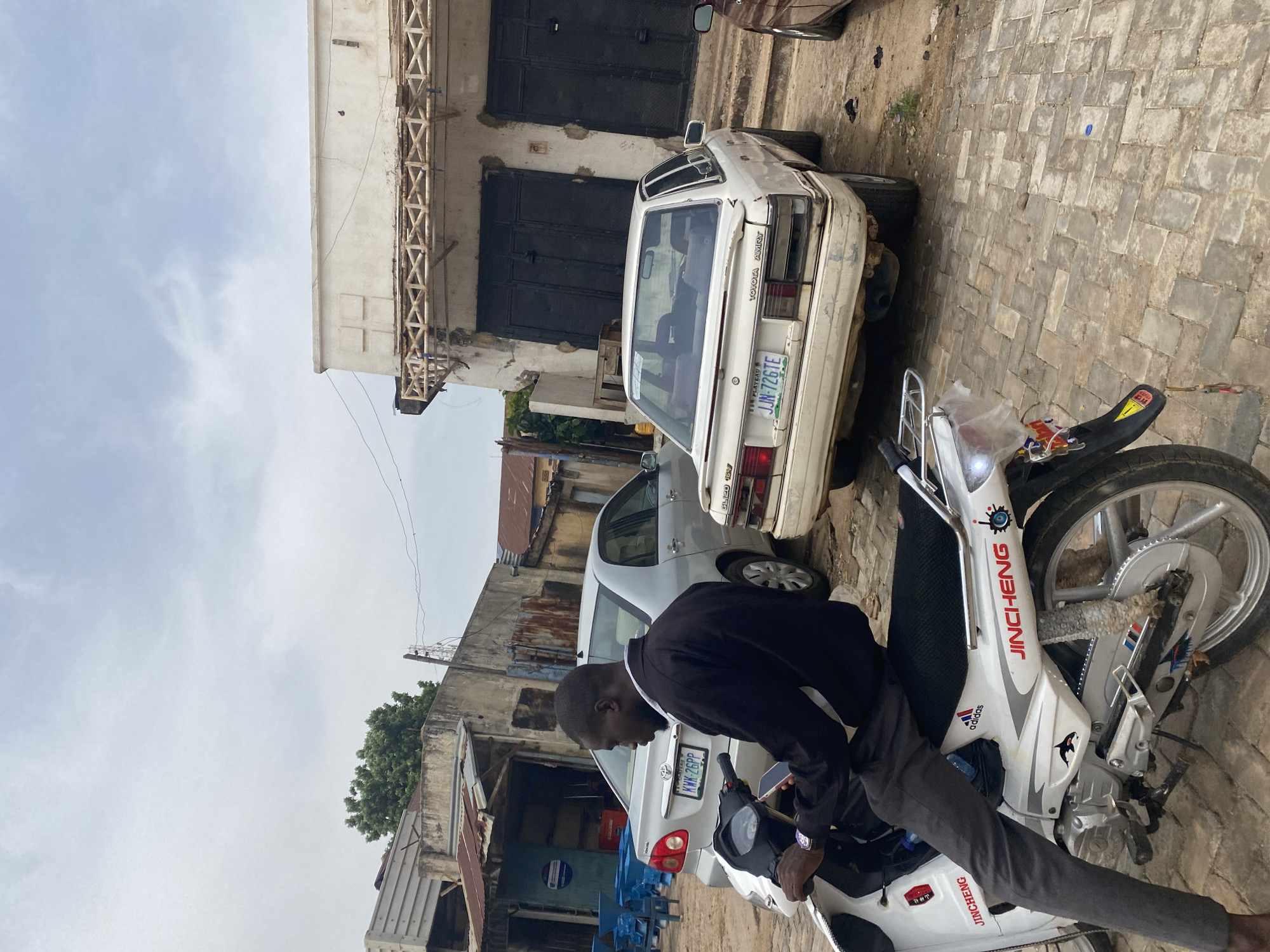 The image depicts a street scene in Jos, Nigeria, with a man riding a white motorcycle in the foreground. The man is wearing a black jacket and grey pants, and he is holding a smartphone in his right hand. The motorcycle has several stickers on it, including "JINCHENG" and an Adidas logo. In the background, there are several cars parked on the street. One car, a white Toyota Camry, has the license plate "JJN-726TE". Another car, a silver Toyota, has the license plate "KWX-26PP". The buildings in the background appear to be older and somewhat run-down. The sky is overcast. The ground is paved with interlocking bricks.