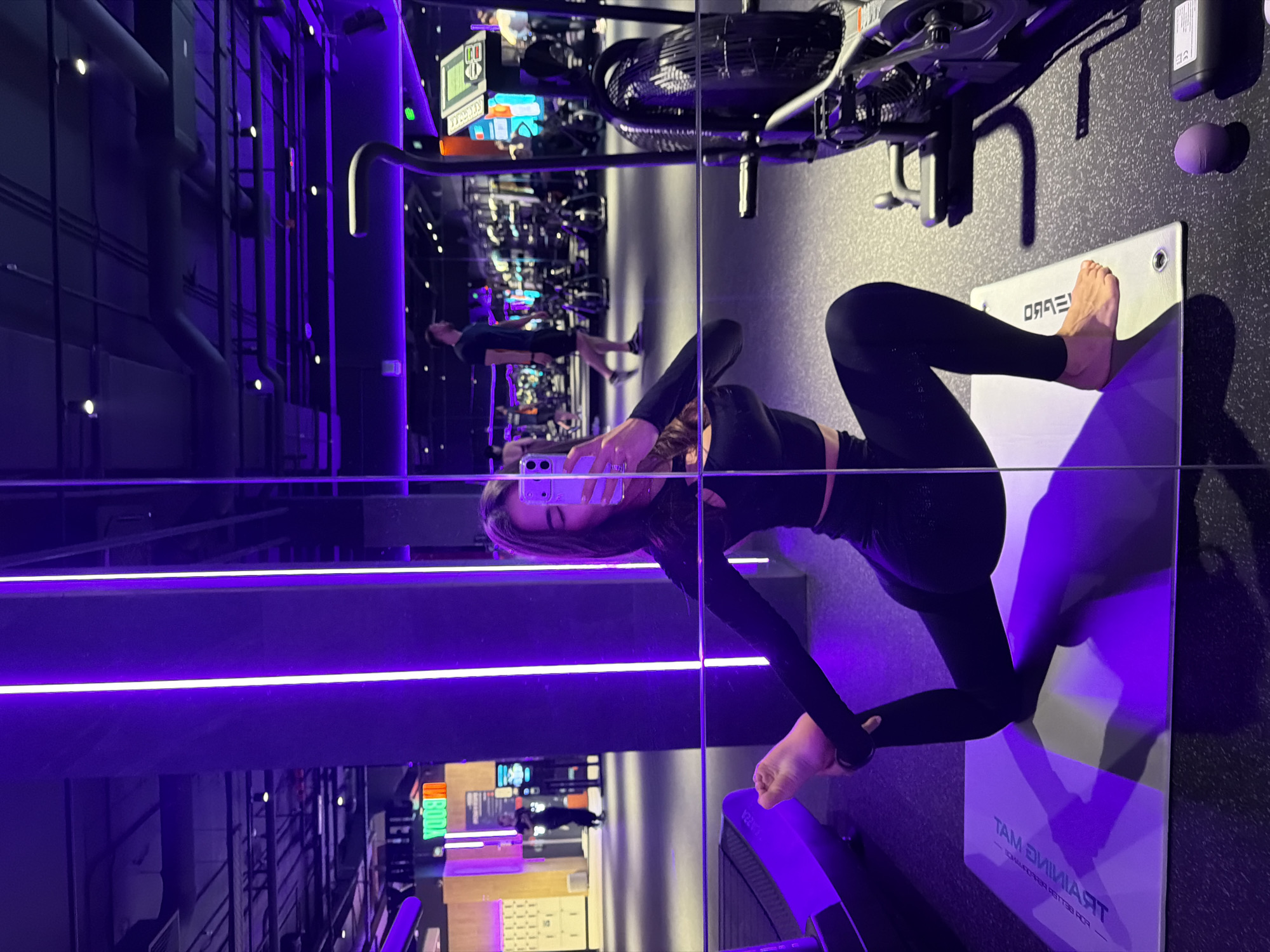 The image displays a modern fitness studio, captured in a sideways orientation where the floor appears as a wall, dominated by dark tones and vibrant purple lighting. The central subject is a woman, reflected in a large mirror that runs vertically through the right side of the frame. She is engaged in taking a selfie, holding a smartphone up to capture her reflection.
The woman is dressed in form-fitting black athletic wear, including a long-sleeved crop top and leggings. She is barefoot, with one foot resting on a white rectangular fitness mat that is positioned on the floor (appearing as a wall in this orientation). The mat features reversed text, clearly reading "TRAINING MAT" and beneath it, "FOR BETTER PERFORMANCE." Her body is posed dynamically, suggesting either a stretch or a moment during an exercise routine.
In the background of the reflection, other gym equipment is visible, including what appears to be a stationary bike or similar machine mounted "upside down" due to the reflection. Another individual, a man, can be seen further back in the reflected gym space. The actual gym structure, visible on the left side of the image, features dark paneled walls with exposed pipes and numerous small, bright lights. Striking purple LED strip lights run horizontally across the scene, contributing to the futuristic and high-tech ambiance of the space. The overall mood is sleek and energetic. The scene is located in a gym in Krasnoyarsk, Russia.