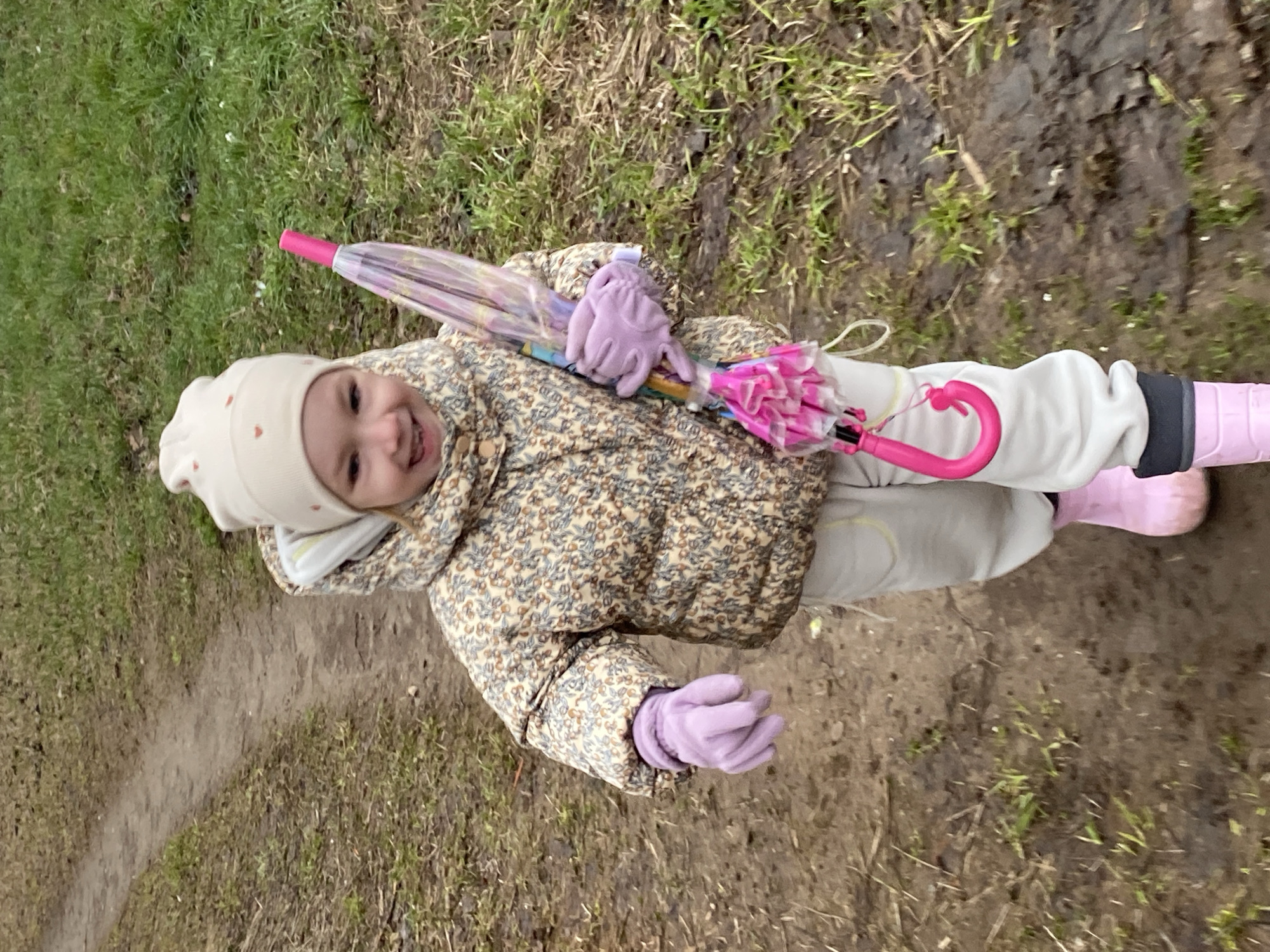 A young child wearing a beige hat with small red hearts, a patterned beige and brown jacket, light gray pants, pink boots, and light purple gloves is holding a closed pink umbrella. The child is standing on a muddy and grassy area, likely a path in a park. The location is possibly Chaikovsky, Russia.
