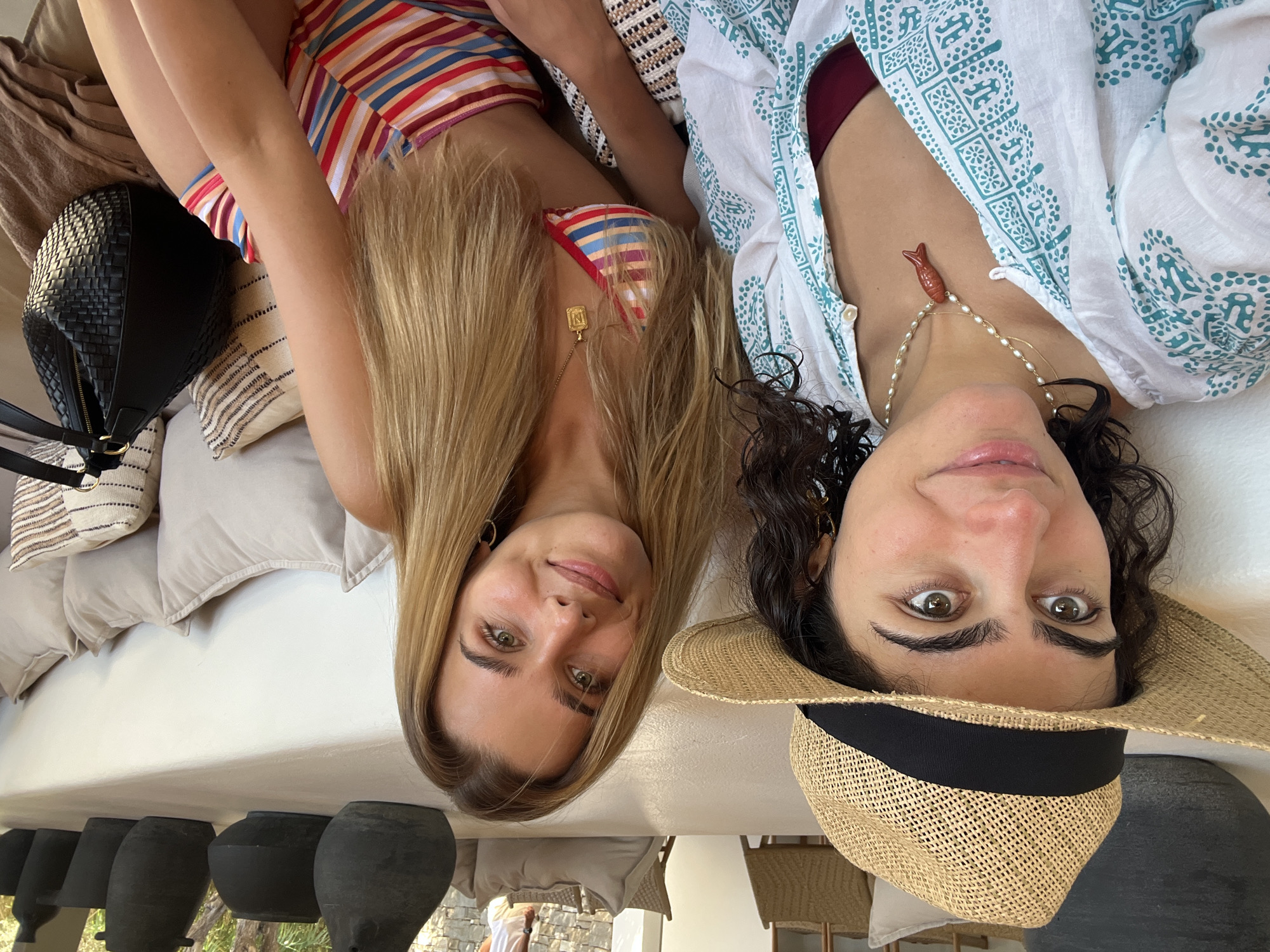 In Naousa, Greece, two young women are seated on a white, built-in couch in an outdoor setting. The woman on the left wears a straw hat, a white and turquoise patterned blouse, and a maroon bikini top. She has dark, curly hair and is wearing a pearl necklace with a red fish pendant. The woman on the right has long, blonde hair and is wearing a colorful striped bikini. She has a gold necklace with a rectangular pendant. Behind them, a long, low table is adorned with several dark, matte ceramic vases. The couch is covered in light-colored cushions, some with patterned covers. A black woven handbag rests on a brown blanket to the right. The background shows glimpses of outdoor seating areas and stone walls, suggesting a relaxed, possibly resort-like environment.