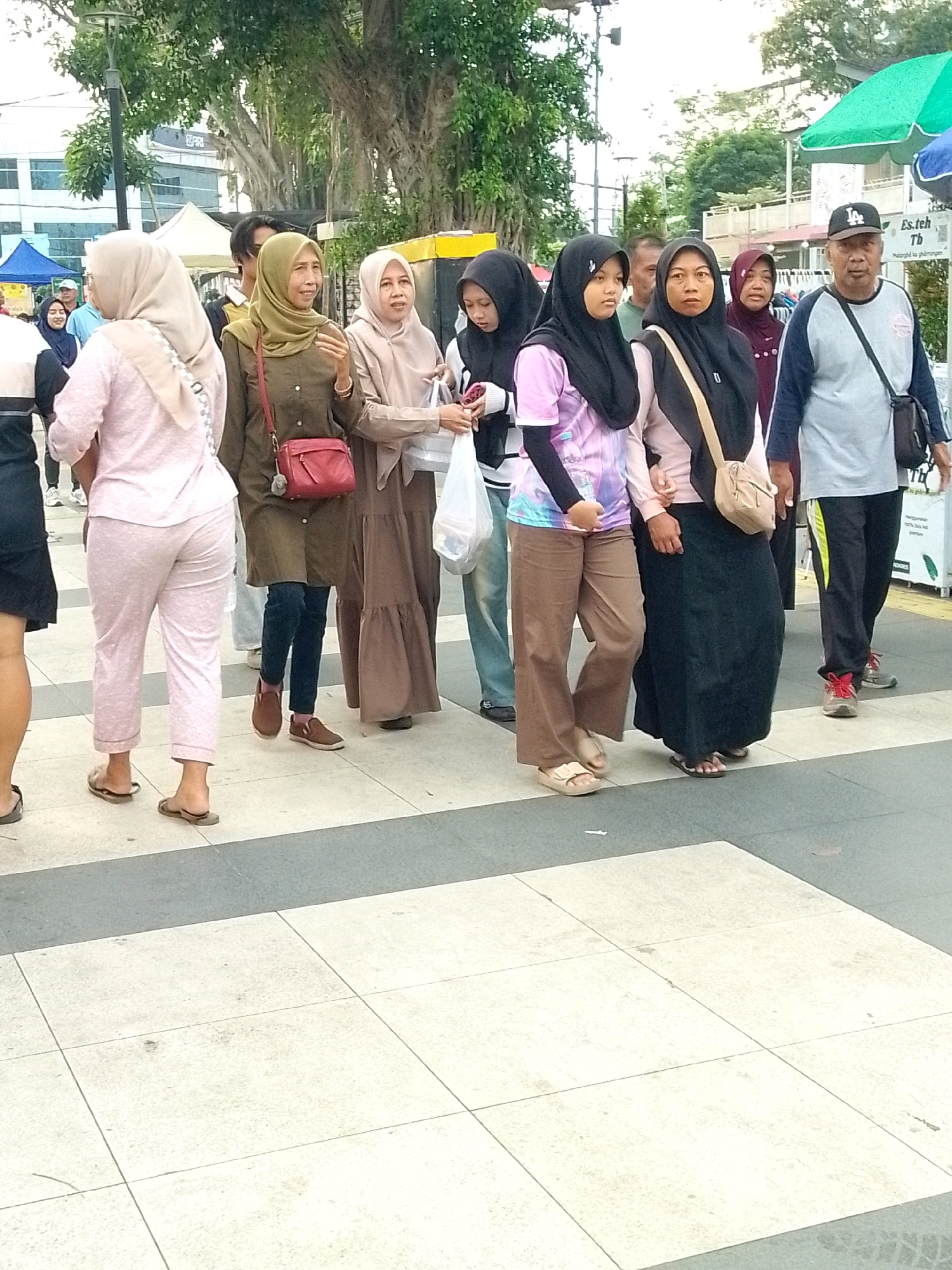 The image captures a group of people walking on a tiled pedestrian area in Jember, Indonesia. In the background, large trees and buildings are visible, suggesting an urban setting, possibly near a commercial or public space. Several individuals are dressed in Islamic modest wear, with women wearing hijabs in various colors and styles. One woman in the foreground wears a light pink hijab and matching pants. To her right, another woman sports a greenish-khaki hijab and a dark green outfit, holding a red purse. Further down the line, a woman in a light brown hijab and a long brown dress is visible, carrying a white plastic bag. Another woman in a black hijab wears a colorful t-shirt and brown pants. A man in a grey and blue long-sleeved shirt with a black bag slung across his chest and a black baseball cap is walking to the right. The scene appears to be a casual daytime outing, possibly during a market day or public event, given the presence of stalls or tents in the background. The ground is paved with light-colored square tiles, interspersed with a grey stripe.