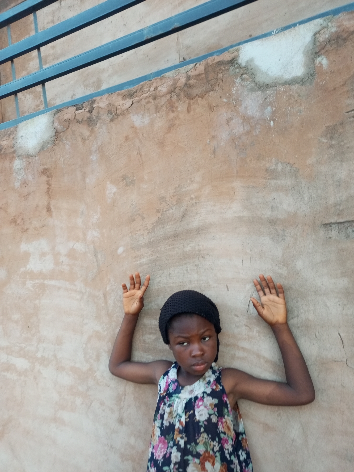 A young girl, appearing to be of African descent, stands with her arms raised and hands against a textured, light brown wall. She is wearing a dark, knitted cap and a sleeveless dress with a floral pattern featuring white, pink, and green flowers on a dark blue background. Her gaze is directed slightly to her right. Above the wall, a section of a blue metal railing is visible, suggesting an outdoor or partially enclosed urban setting. The wall exhibits signs of age and wear, with patches of lighter material and some cracking. The lighting suggests it is daytime. The scene is set in Tempure, Nigeria.