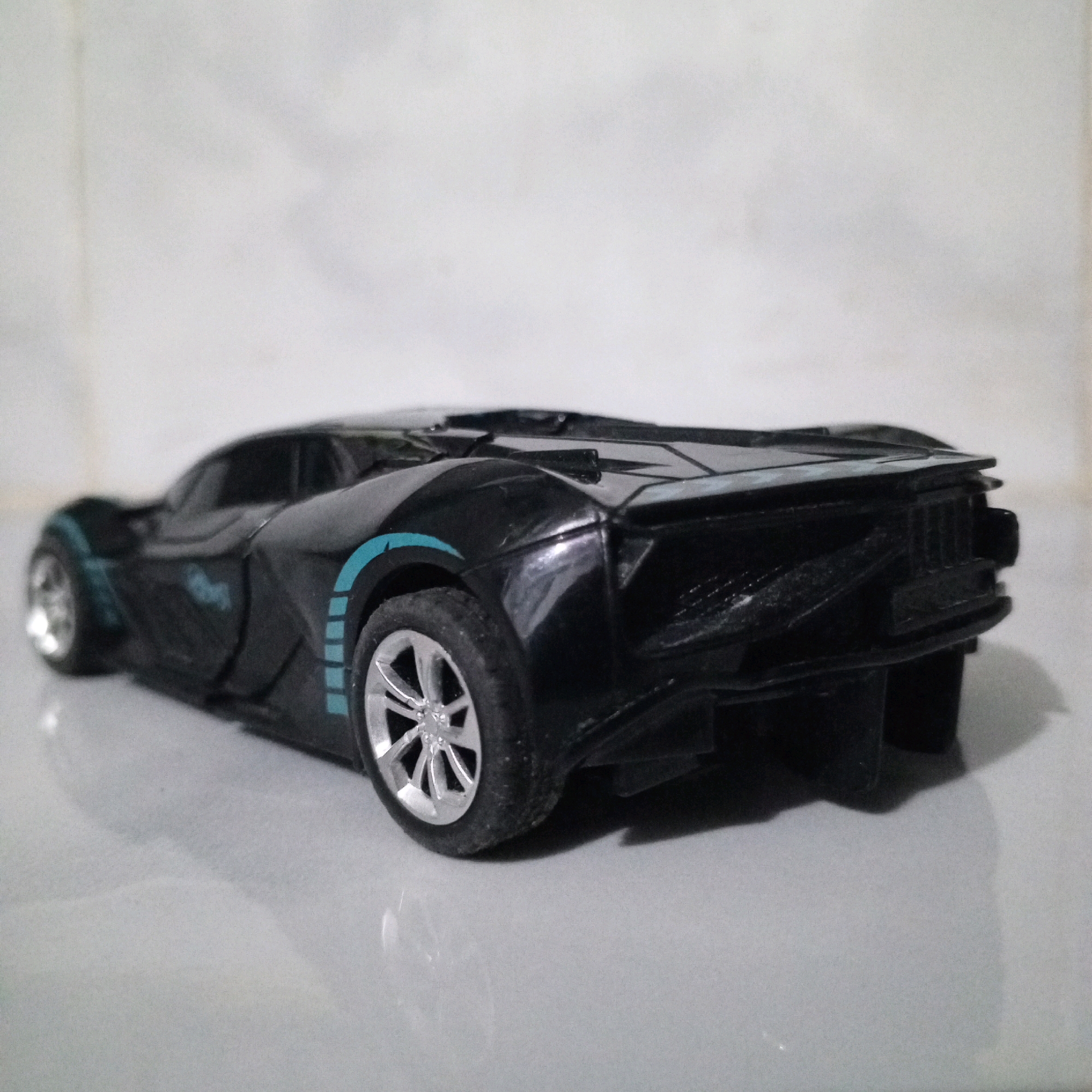 A black toy sports car is the sole subject in this image, positioned on a reflective gray surface. The car is viewed from a rear-three-quarter angle, with its right side predominantly in view. It features prominent teal racing stripes on its rear fender and a similarly colored accent on the front fender. The car's bodywork is angular and futuristic. The background is a lightly textured, off-white surface. The image is an indoor photograph, with lighting that casts a shadow from the car onto the reflective surface below. There is no indication of the city of Kolursari, Indonesia, or any other specific location within the scene.
