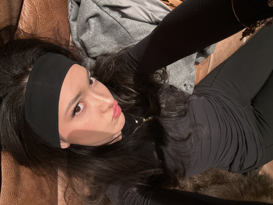 A woman with long, dark hair and a black headband is lying down. She's wearing a black top and leggings, posing for a selfie with pursed lips. A gray garment is visible behind her head. She is wearing a delicate necklace. The background appears to be a brown leather couch. The lighting is subdued, casting soft shadows on her face.