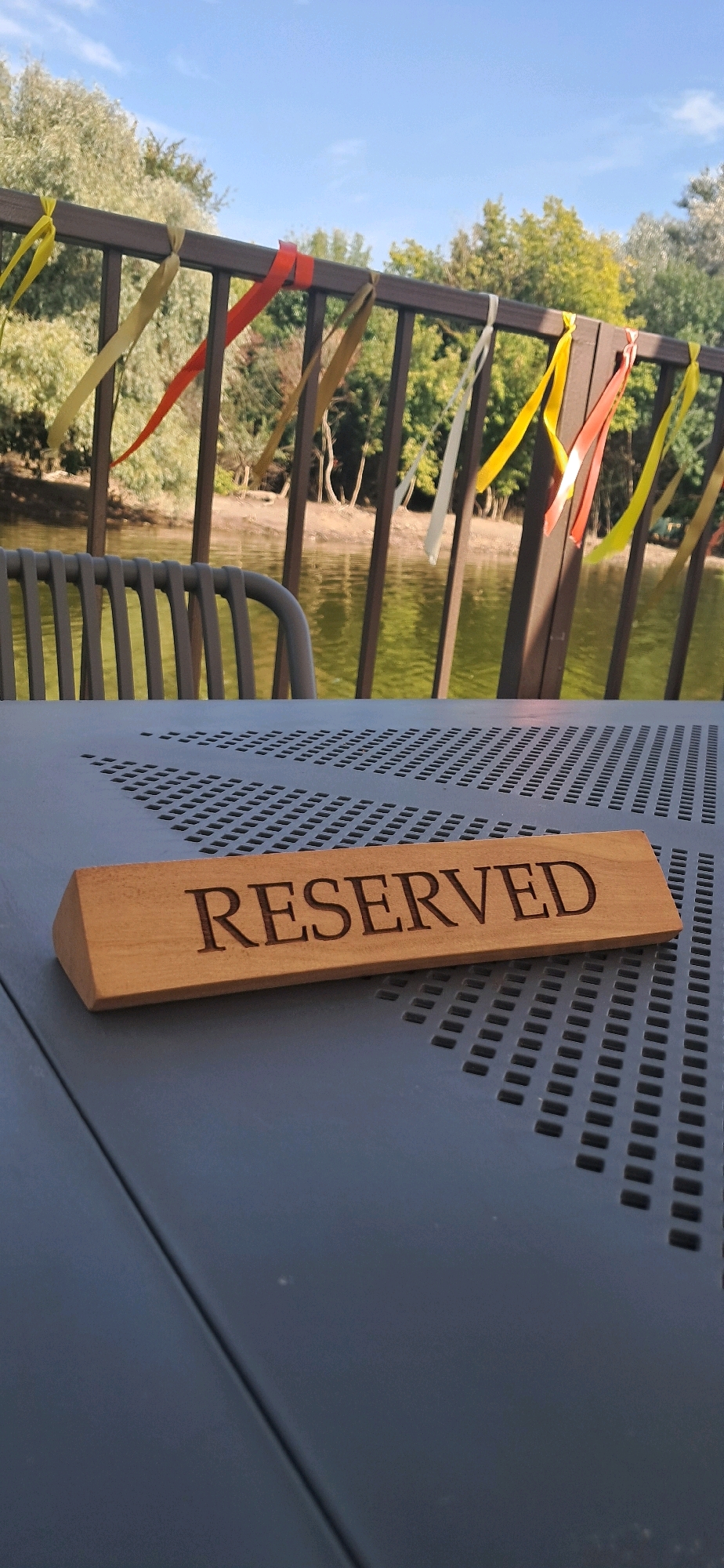 The image depicts an outdoor scene, likely in Chykalivka, Ukraine, with a focus on a wooden "RESERVED" sign placed on a dark, perforated surface, possibly a table. In the background, a body of water is visible, reflecting the light and bordered by trees. A dark railing with colorful ribbons attached to it runs along the edge of the water. A metal chair is partially visible in the midground, also facing the water. The sky above is clear and blue. The scene suggests a relaxed outdoor setting, perhaps a restaurant or cafe by the water.