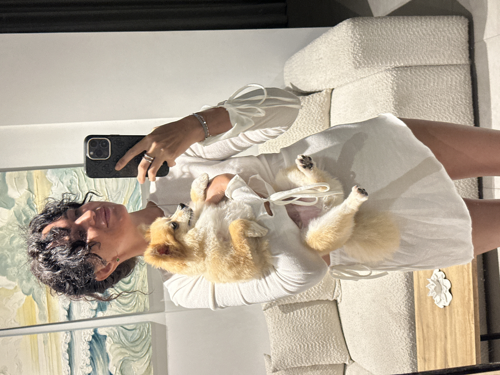A woman with dark, curly hair is holding a small, fluffy dog. She is wearing a white, long-sleeved dress with ties on the sleeves. The woman is holding her phone in her right hand, taking a selfie. She is wearing rings on her left hand and a bracelet on her right wrist. The dog is light brown and appears to be a Pomeranian.
In the background, there is a cream-colored couch with textured pillows. To the left of the couch, there are two framed artworks depicting ocean waves. A wooden coffee table is in the foreground, with a white floral-shaped object on it. The scene appears to be indoors, possibly in a living room or lounge area.
The location of this image is not specified.