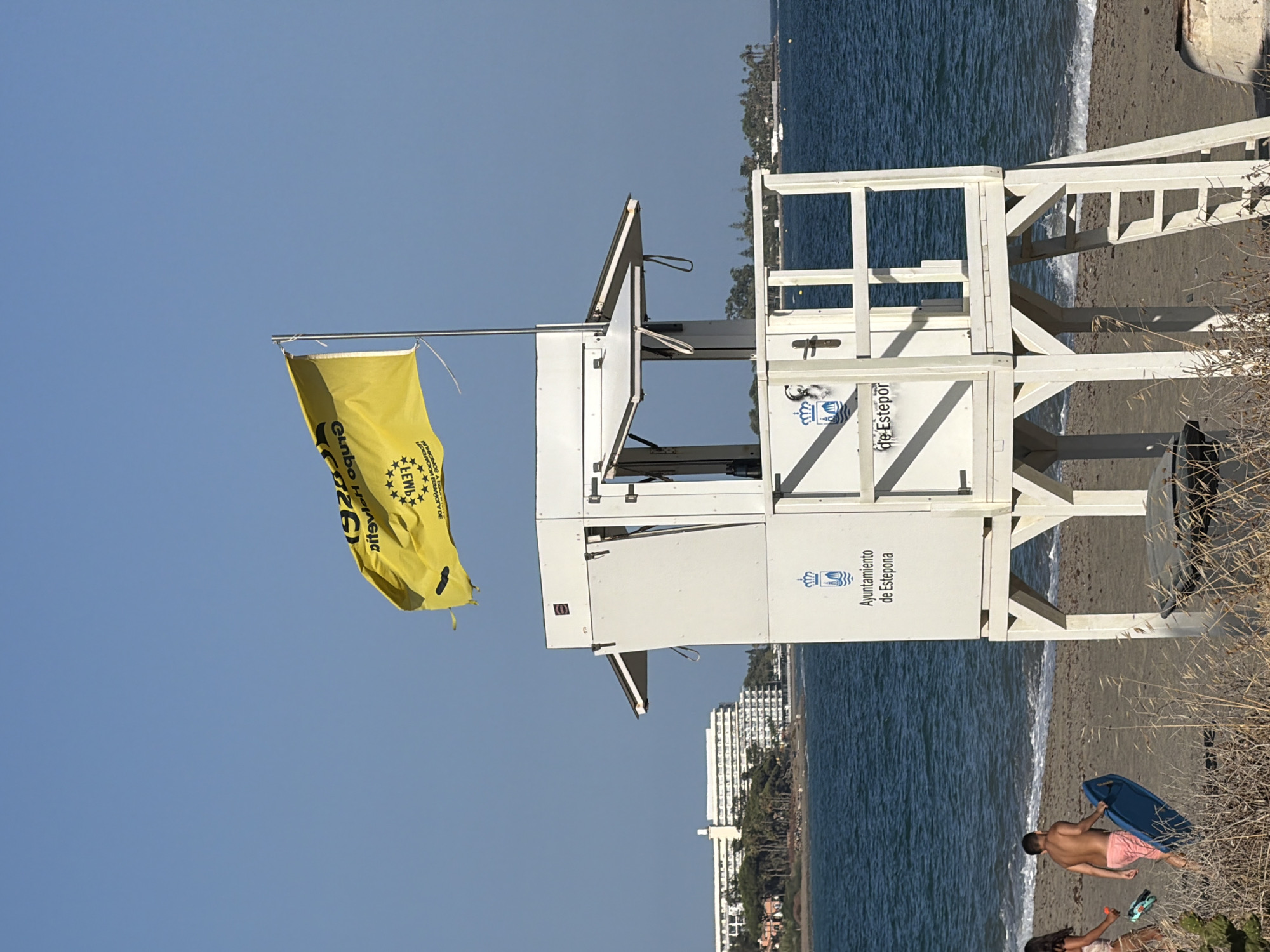 A bright yellow flag with black text and stars is unfurled on a flagpole against a clear blue sky. Below the flag is a white lifeguard tower with a visible ladder. The tower has text and a crest on its side, reading "Ayuntamiento de Estepona" and "Km0 Estepona". A body of water, likely the sea, is visible behind the tower, with a sandy beach in the foreground. Two people, a woman and a boy, are walking on the beach near the water's edge. The boy is carrying a blue boogie board. In the background, a large white building complex with many windows is visible on a hill overlooking the sea, surrounded by trees.
