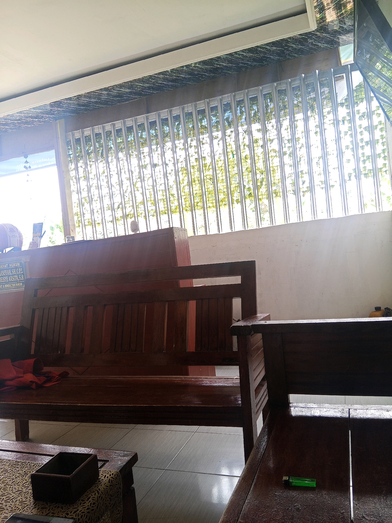 The image shows a simple waiting area, likely at a government office or small business in Lubuklinggau, Indonesia. The primary subjects are two dark wooden benches, one in the foreground and one in the background. The bench in the background appears to be a three-seater with a dark red cloth draped over one end. On the wall behind it, there is a sign with text in Indonesian that seems to list names and legal titles. A framed object and a white circular object are also visible on the wall. The bench in the foreground is a two-seater, and on its surface, there is a green disposable lighter. The floor is tiled with light gray square tiles. Above the benches, there is a window covered by a metal grate, allowing natural light to filter through, illuminating a pattern of foliage. The ceiling is white with a decorative textured panel. The overall impression is one of a functional, no-frills waiting space.