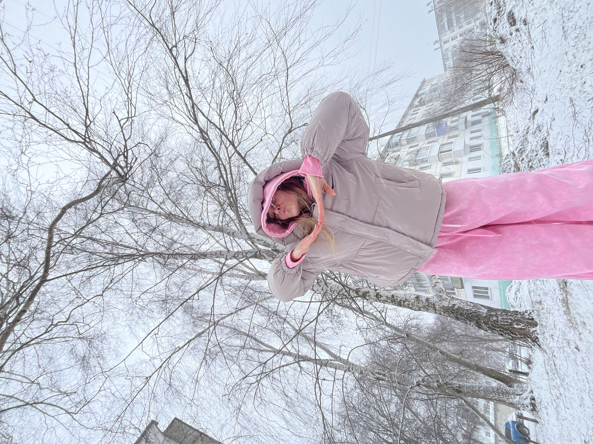 The image shows a woman outdoors in winter. She is wearing a pink tracksuit and a light gray puffer jacket with the hood up, and she is standing in a snowy environment with bare trees surrounding her. The woman's face is visible within the hood, and she has her hands up near her face. Behind the woman and trees, a tall building with numerous windows is visible. The location of the image is identified as the city of Chaikovsky, Russia.