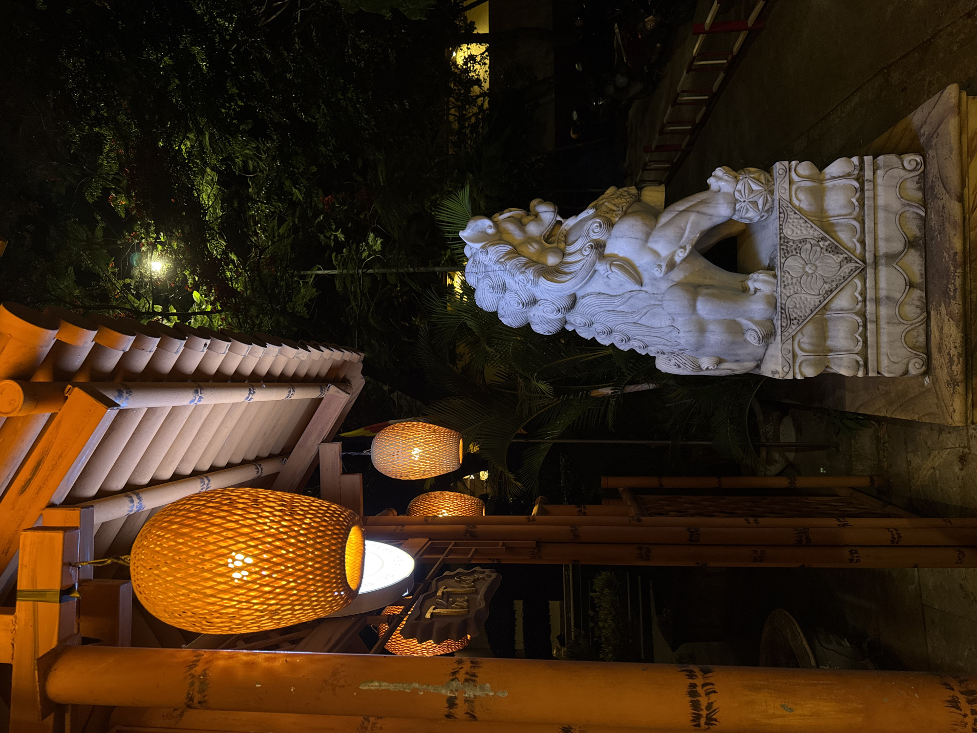 The image depicts the exterior of a traditional Vietnamese establishment at night, likely a restaurant or shop, located in Đà Nẵng, Vietnam. The foreground features a large, intricately carved white stone lion statue, a common motif in East Asian architecture, placed on a stone pedestal. To the left, an orange-painted bamboo pillar stands prominently, with a woven bamboo lantern hanging from an orange wooden beam above. More similar lanterns are visible, casting a warm, orange glow. A wooden sign with Vietnamese script, appearing to read "Tri Thu," is mounted on the bamboo structure. Behind the lion statue, a pathway is visible, bordered by a red and white railing. Lush green foliage and trees are present in the background, illuminated by a distant street light. The overall ambiance is atmospheric and traditional, suggesting an authentic cultural experience.